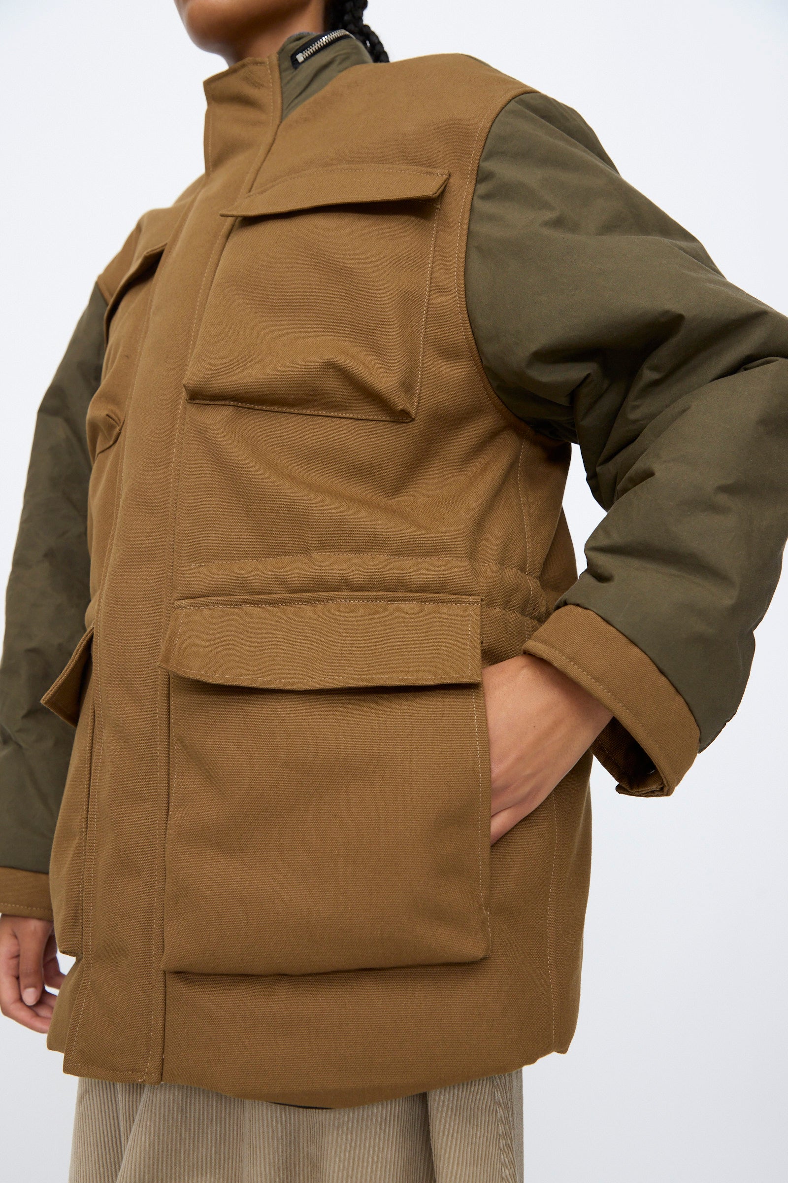 Person wearing the Kassl Canvas Wax Field Jacket in Almond and Oil Black, styled with a brown utility vest, standing against a plain light background.