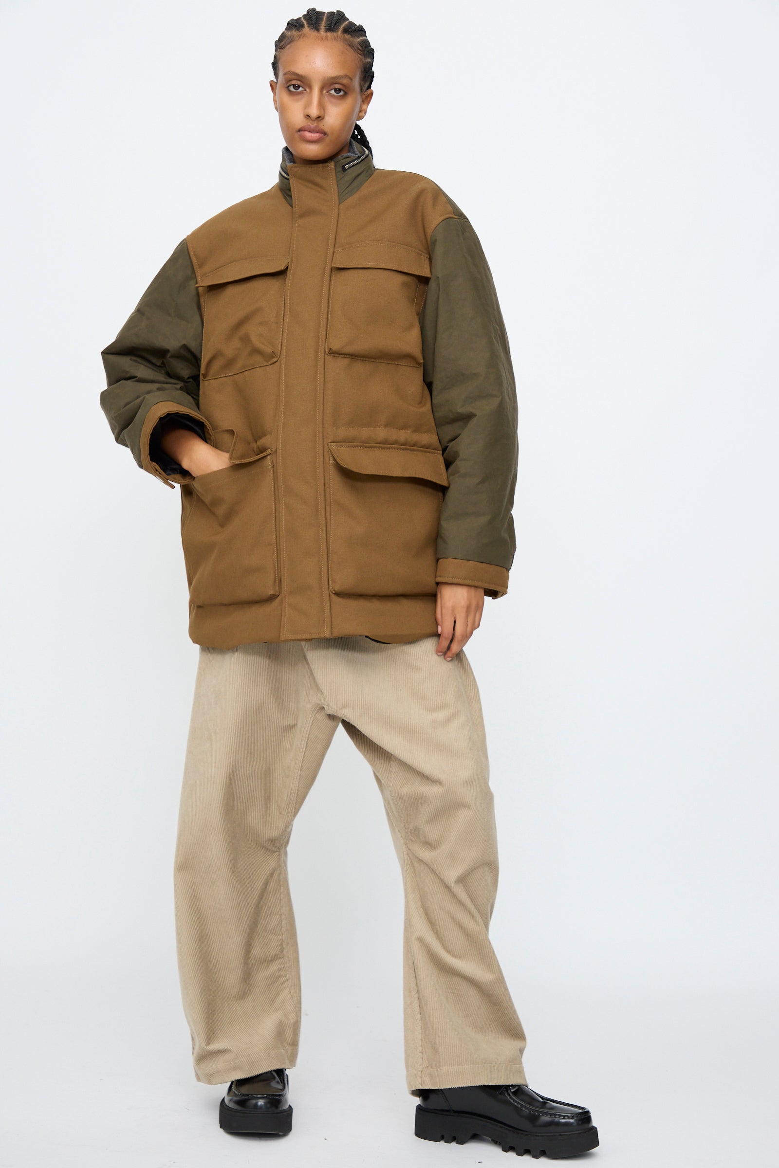 A person wears the Kassl Canvas Wax Field Jacket in Almond and Oil Black with beige wide-leg pants and black shoes, standing against a plain white background.