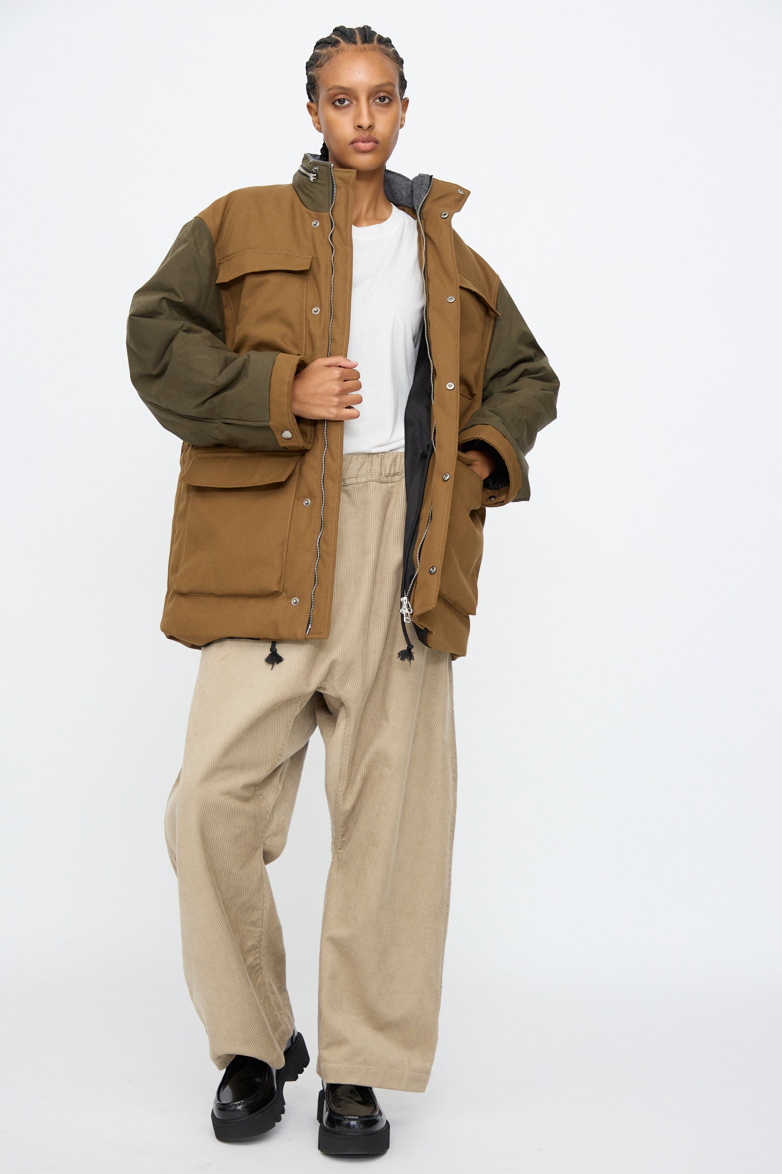 Person standing against a white background, wearing the Kassl Canvas Wax Field Jacket in Almond and Oil Black with an adjustable waist, a white T-shirt, wide beige pants, and black shoes.