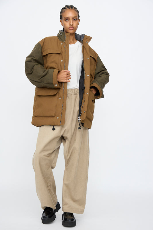 Person standing against a white background, wearing the Kassl Canvas Wax Field Jacket in Almond and Oil Black with an adjustable waist, a white T-shirt, wide beige pants, and black shoes.