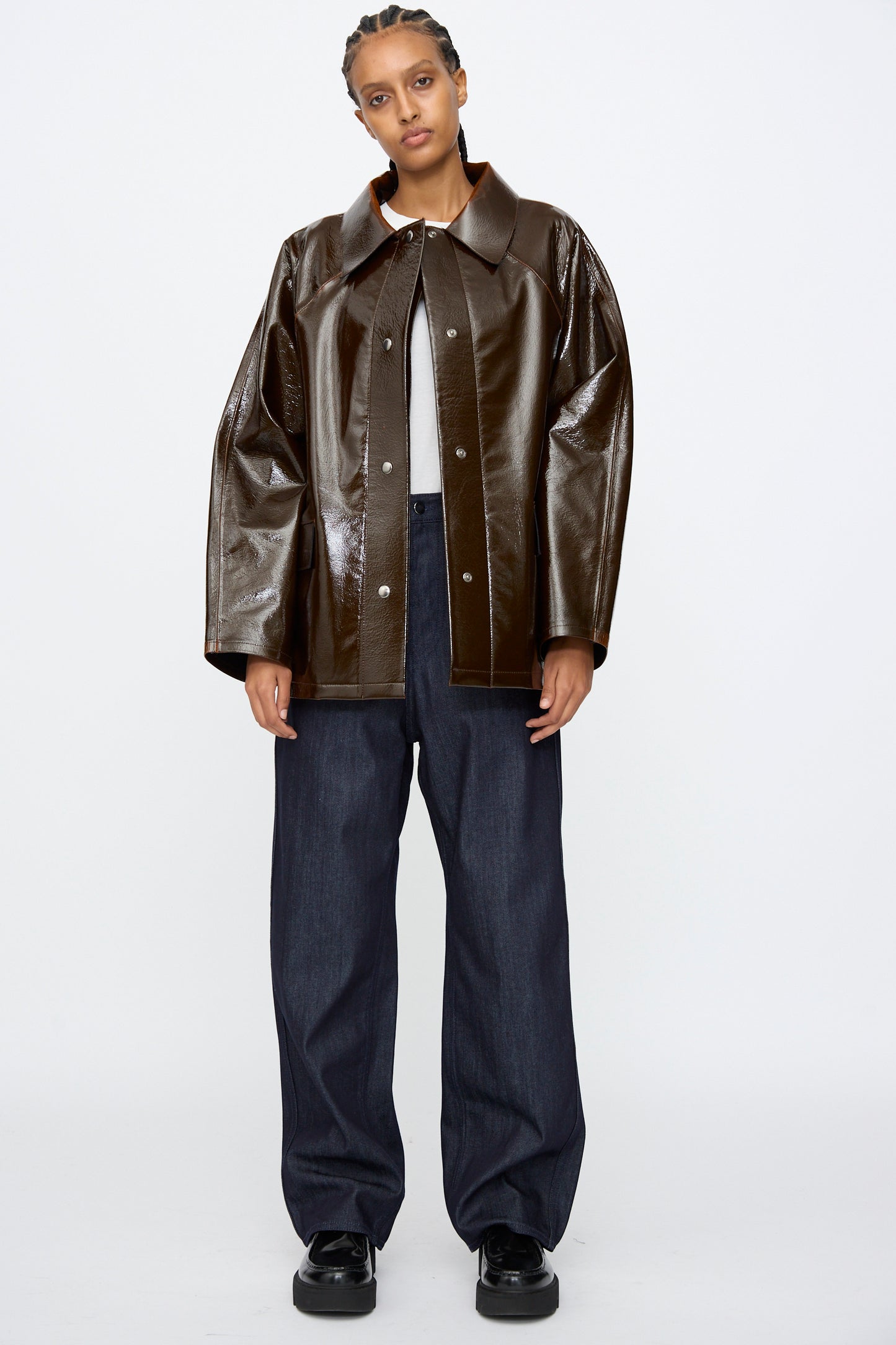 A person stands facing forward in the Kassl Laquer Wool Original Hip Jacket in Honey, featuring a relaxed fit, paired with a gray shirt, wide-leg dark jeans, and black shoes against a white background.