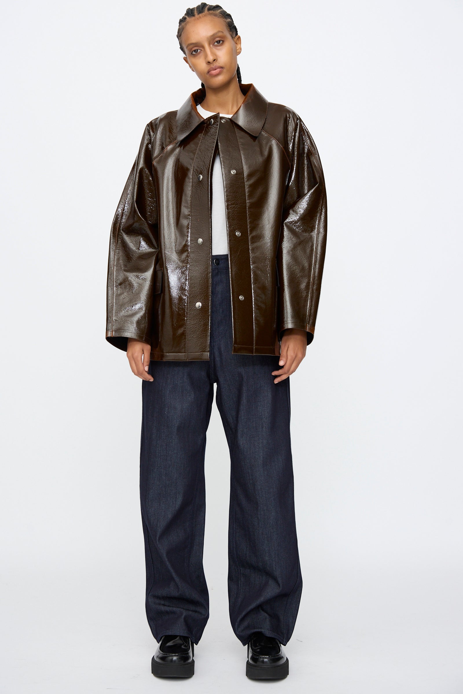 A person stands facing forward in the Kassl Laquer Wool Original Hip Jacket in Honey, featuring a relaxed fit, paired with a gray shirt, wide-leg dark jeans, and black shoes against a white background.