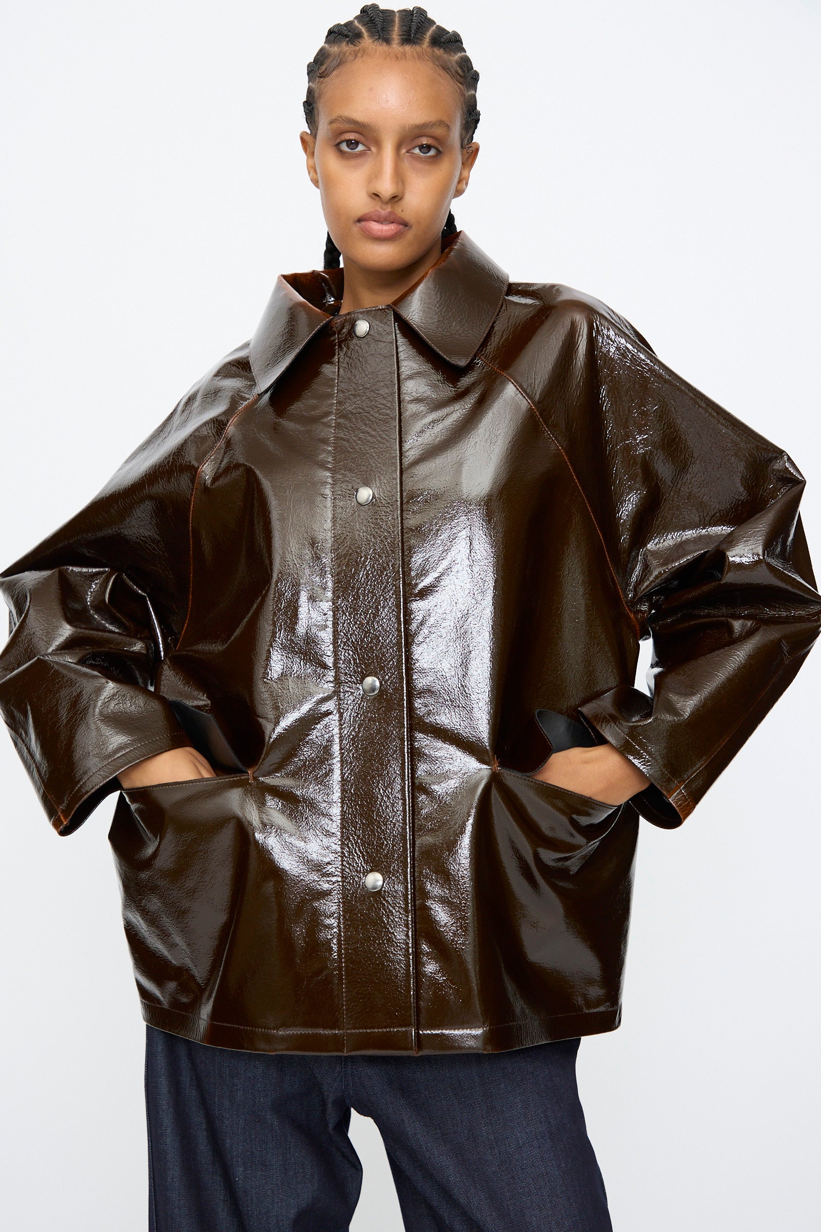 A person stands with hands in pockets, wearing the Kassl Laquer Wool Original Hip Jacket in Honey—a relaxed-fit piece with a shiny finish, oversized button-up design, wide sleeves, and a large collar.