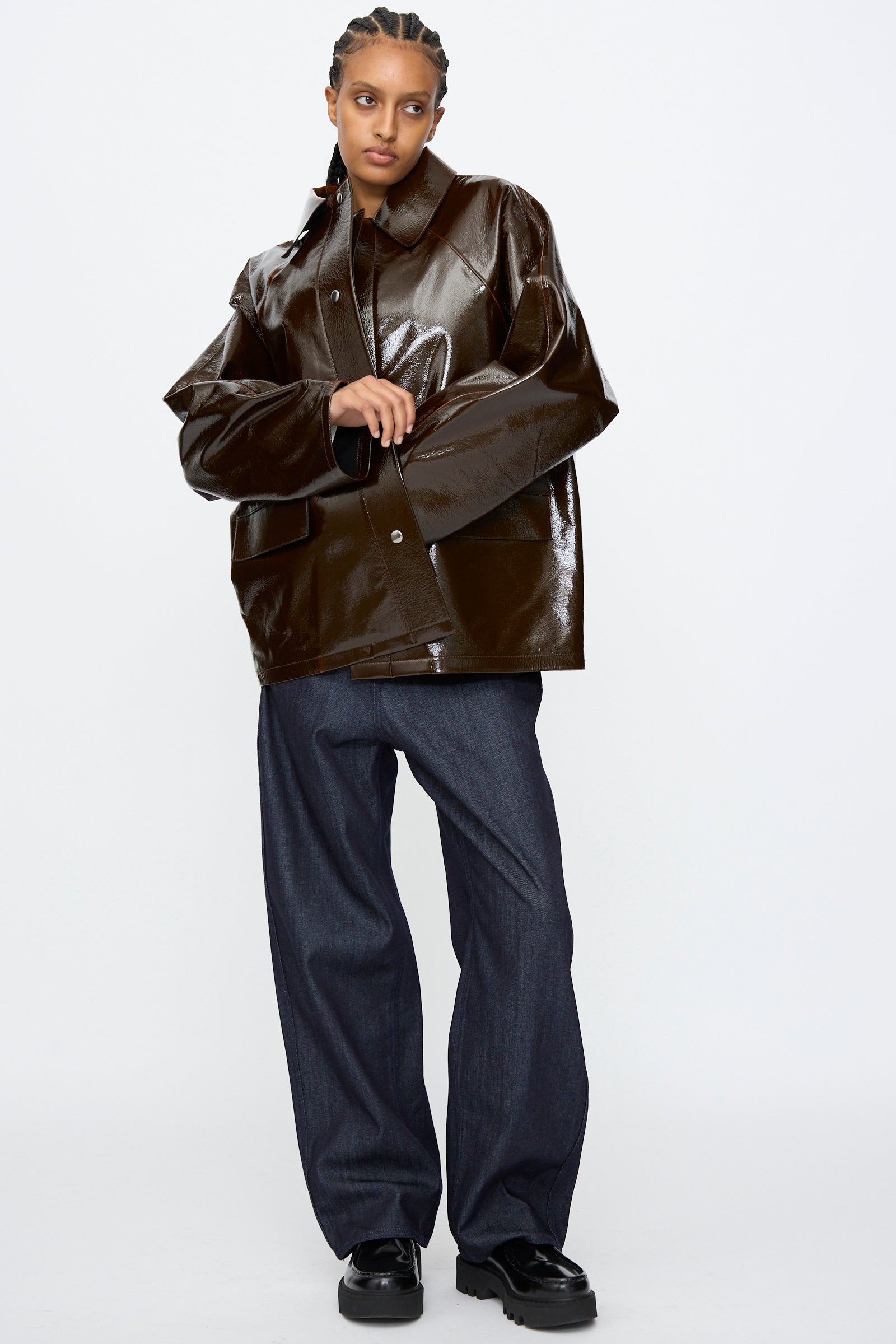 Person stands against a plain background, wearing the Kassl Laquer Wool Original Hip Jacket in Honey, paired with relaxed fit dark pants and black shoes, with braided hair.