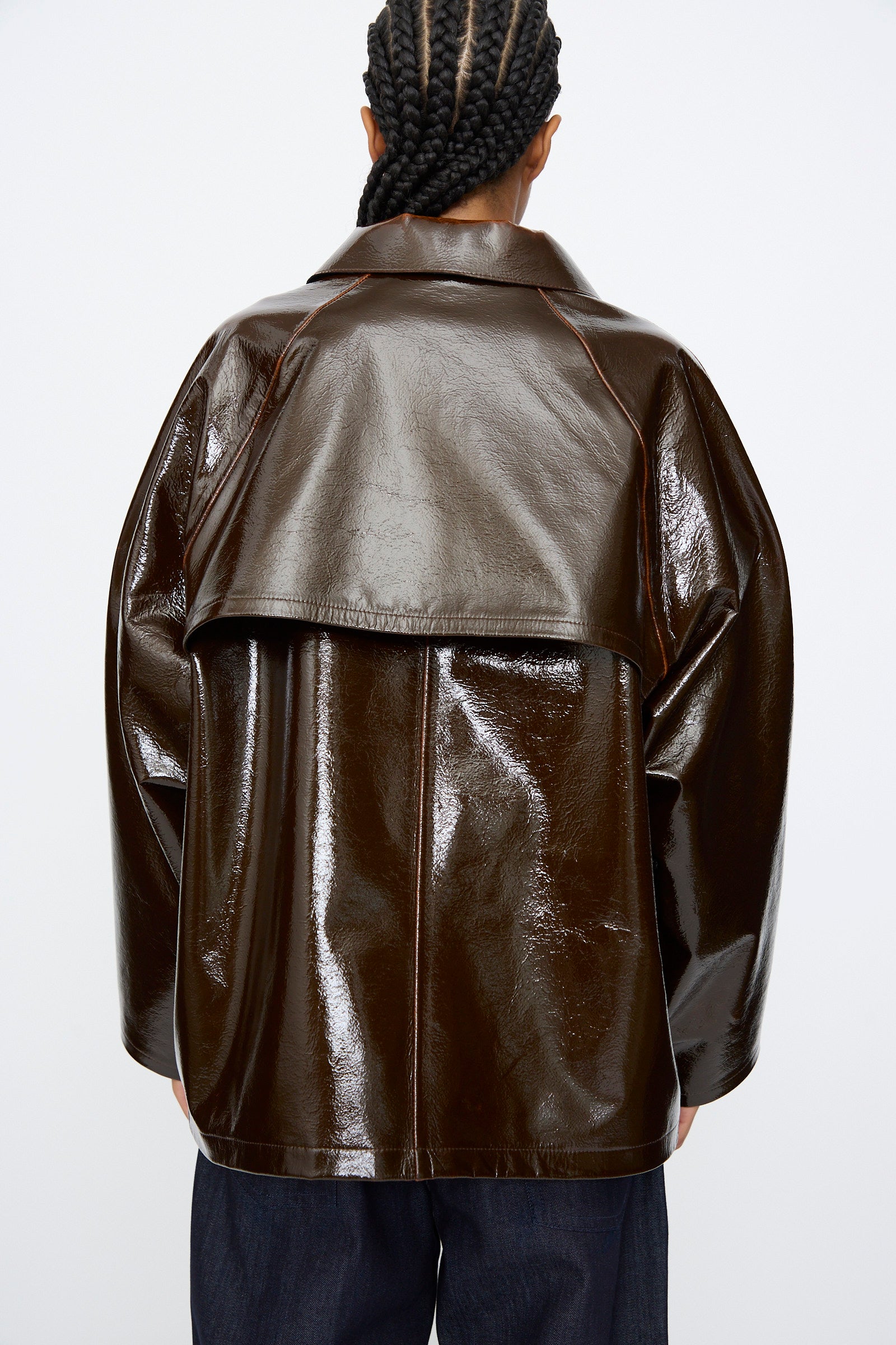 A person with braided hair wears the Kassl Laquer Wool Original Hip Jacket in Honey, featuring a shiny finish and relaxed fit, photographed from the back against a plain light background.