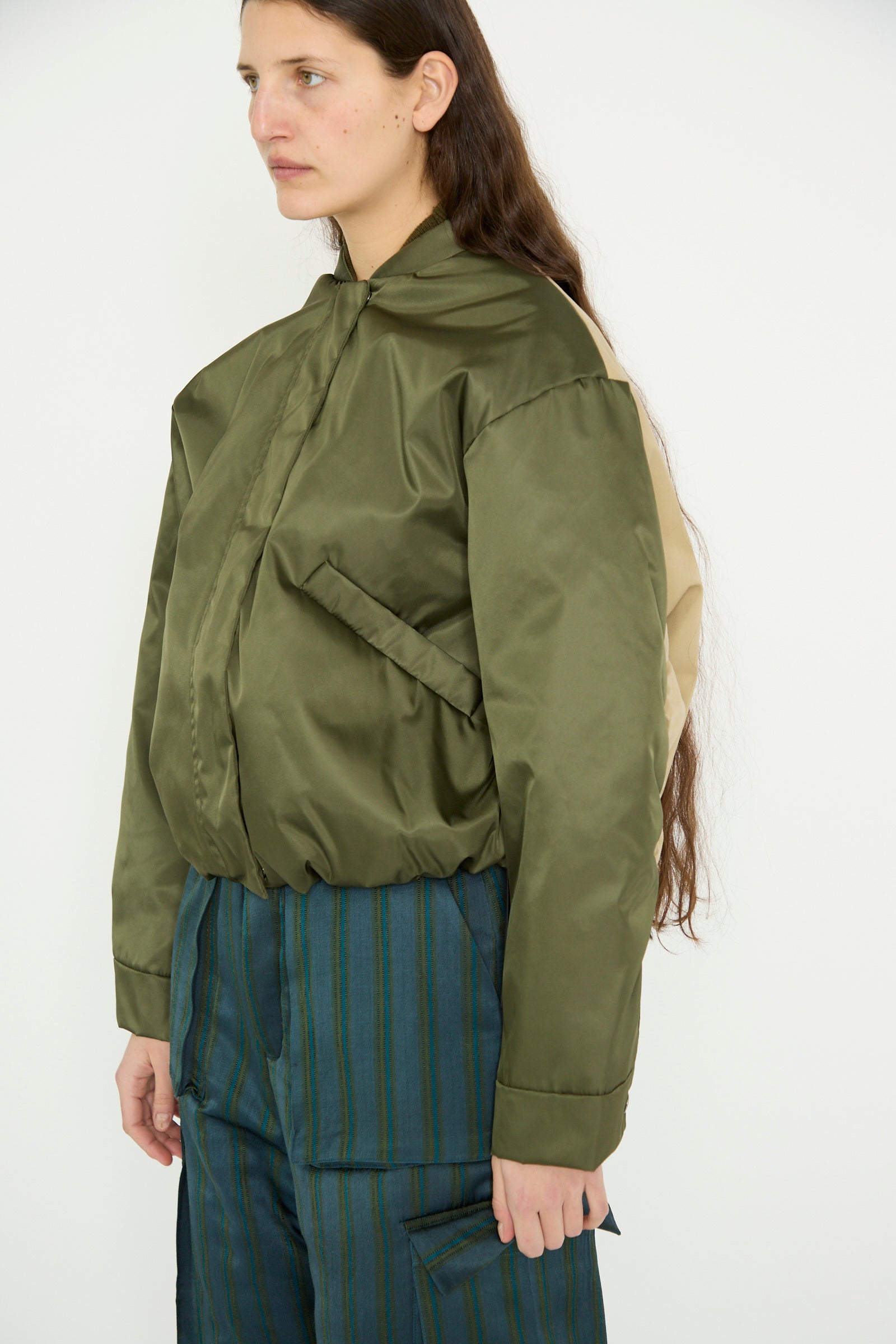 A person with long brown hair wears Kassl's Nylon Bomber Short in Khaki and Trench Beige against a plain background, their coat featuring an adjustable waist for comfort.