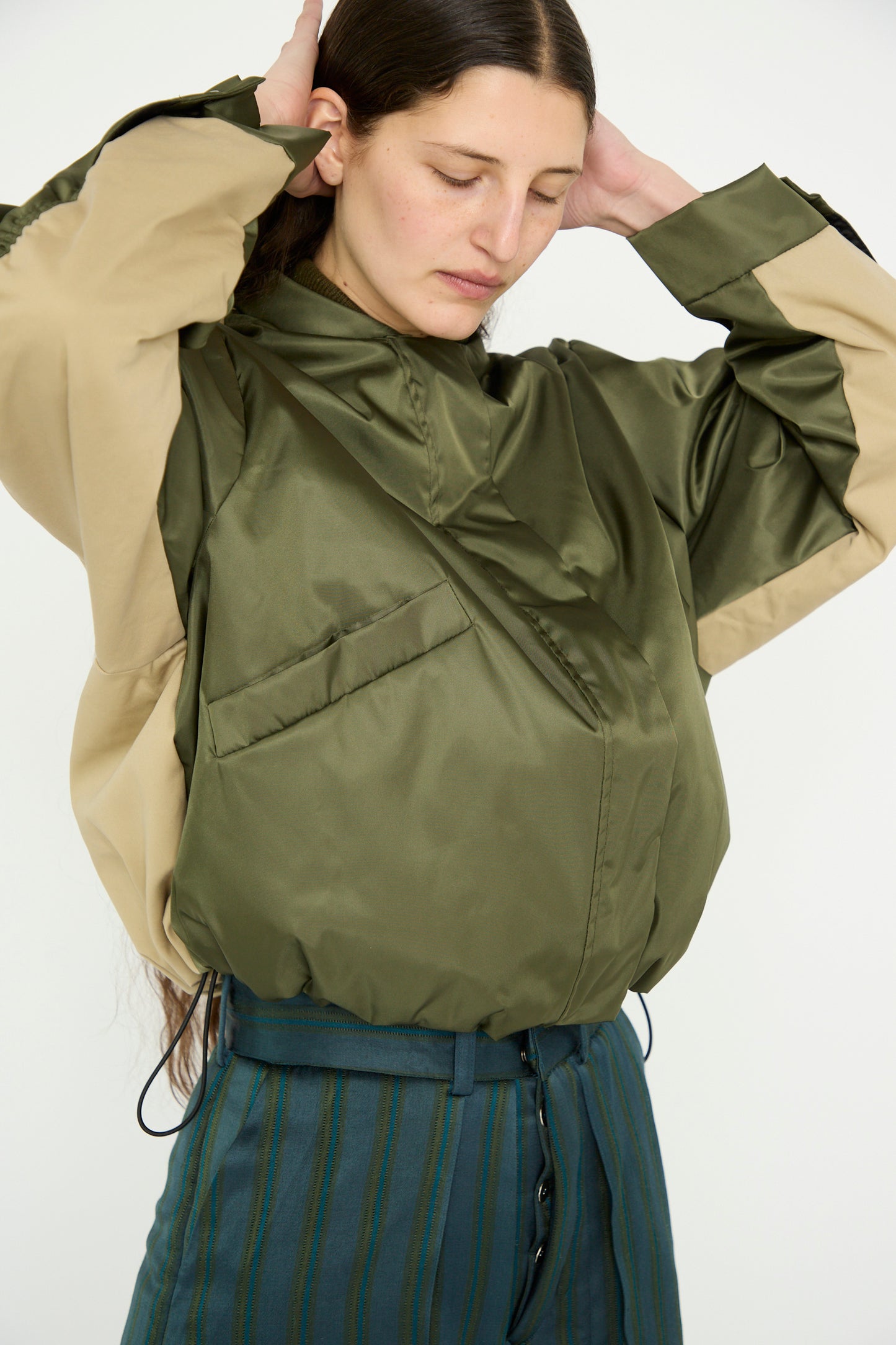 A person adjusts their hair while wearing the Kassl Nylon Bomber Short, a technical jacket in khaki and trench beige, paired with high-waisted striped green and blue pants against a neutral background.