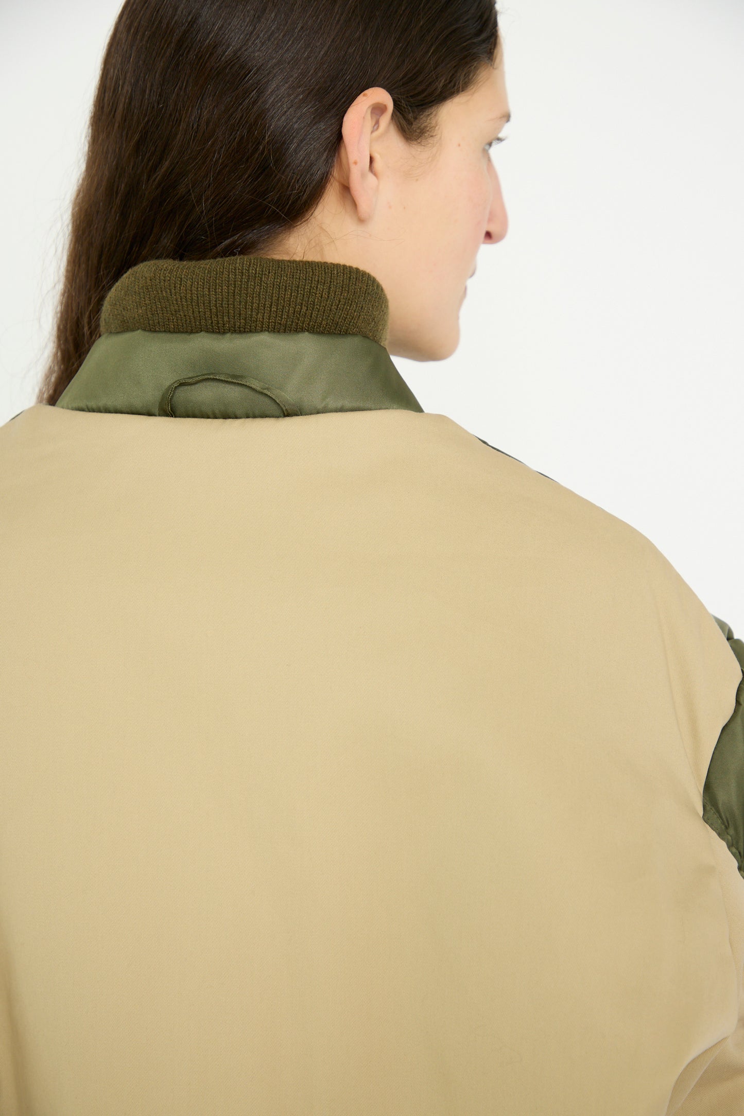 Rear view of a person with long dark hair in a Kassl Nylon Bomber Short jacket in Khaki and Trench Beige, featuring a dark green collar and adjustable waist.