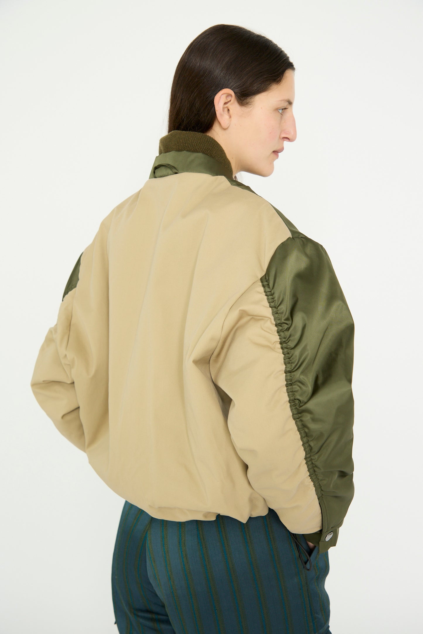 A person is seen in a side profile against a white background, wearing Kassl's Nylon Bomber Short jacket in Trench Beige and Khaki, featuring green sleeves and an adjustable waist.