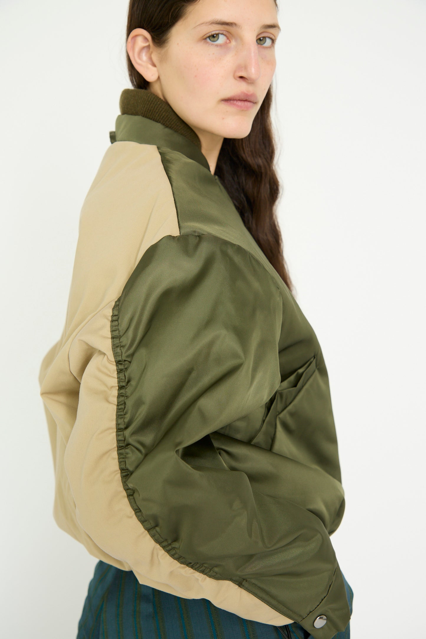 A person with long hair wears a Kassl Nylon Bomber Short in Khaki and Trench Beige, glancing over their shoulder against a plain background.