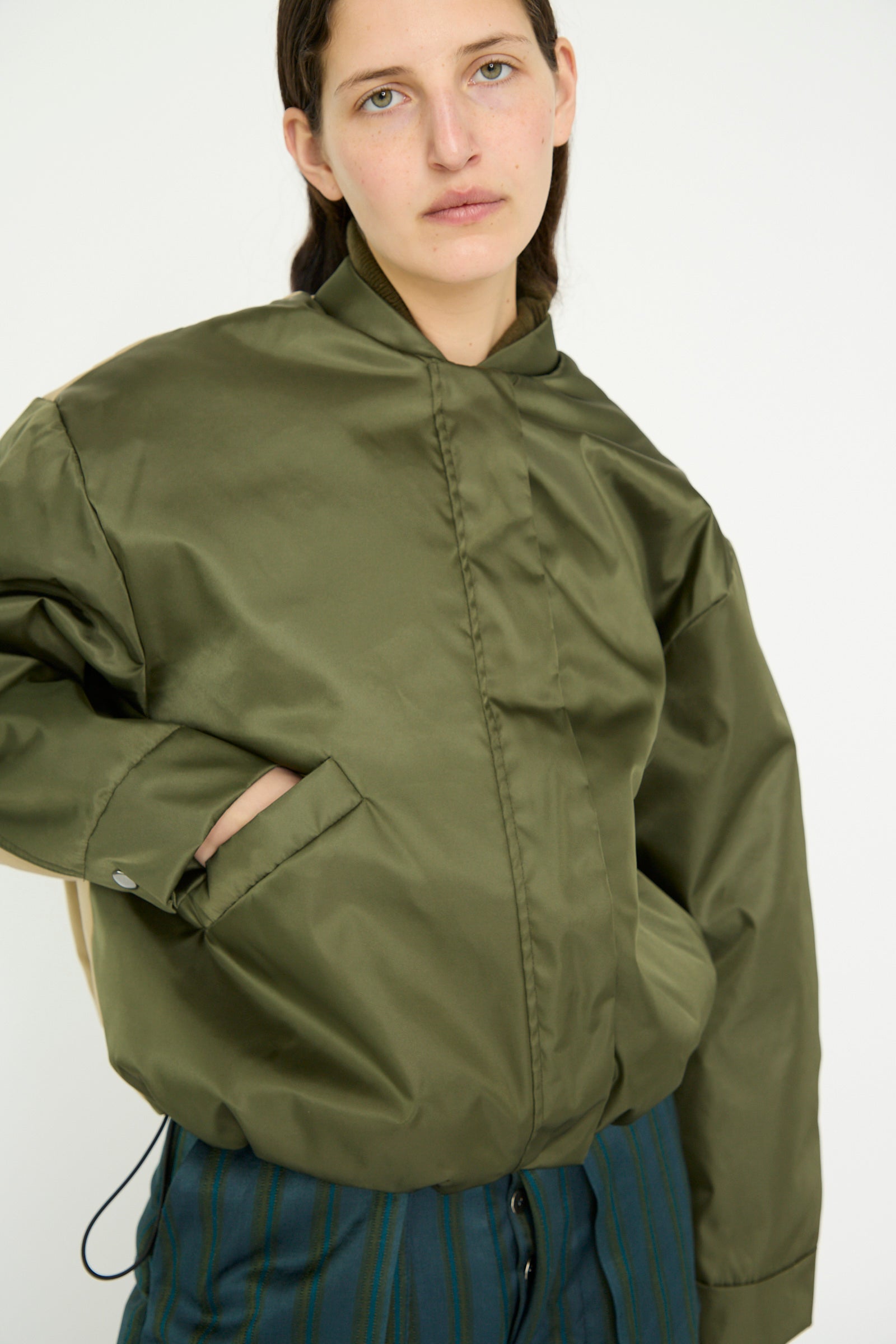 A person wearing the Kassl Nylon Bomber Short in Khaki and Trench Beige with an adjustable waist, hands in pockets, stands against a white background.