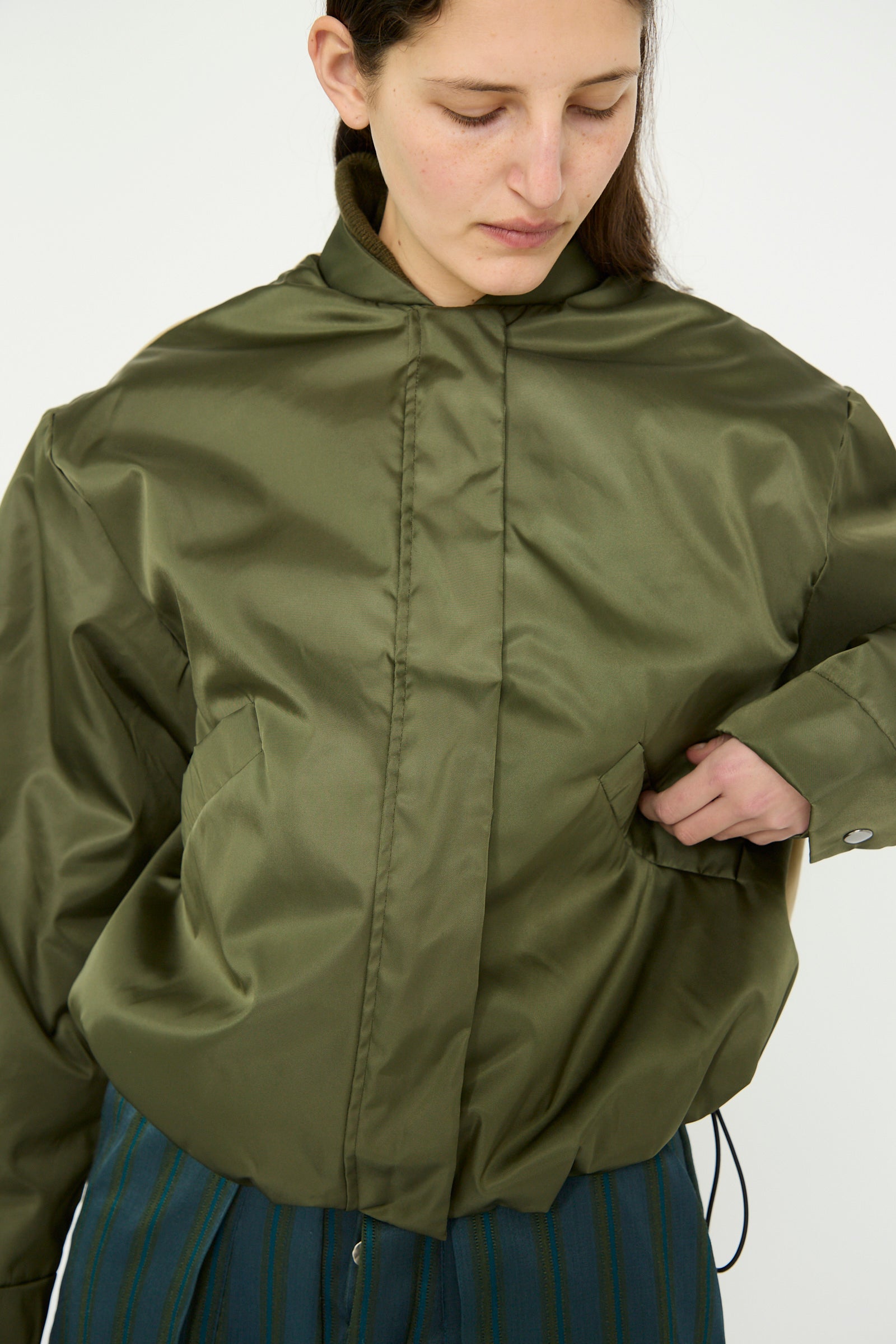 A person in Kassl's Nylon Bomber Short in khaki and trench beige, with a green, oversized design, stands with one hand in their pocket, gazing down.