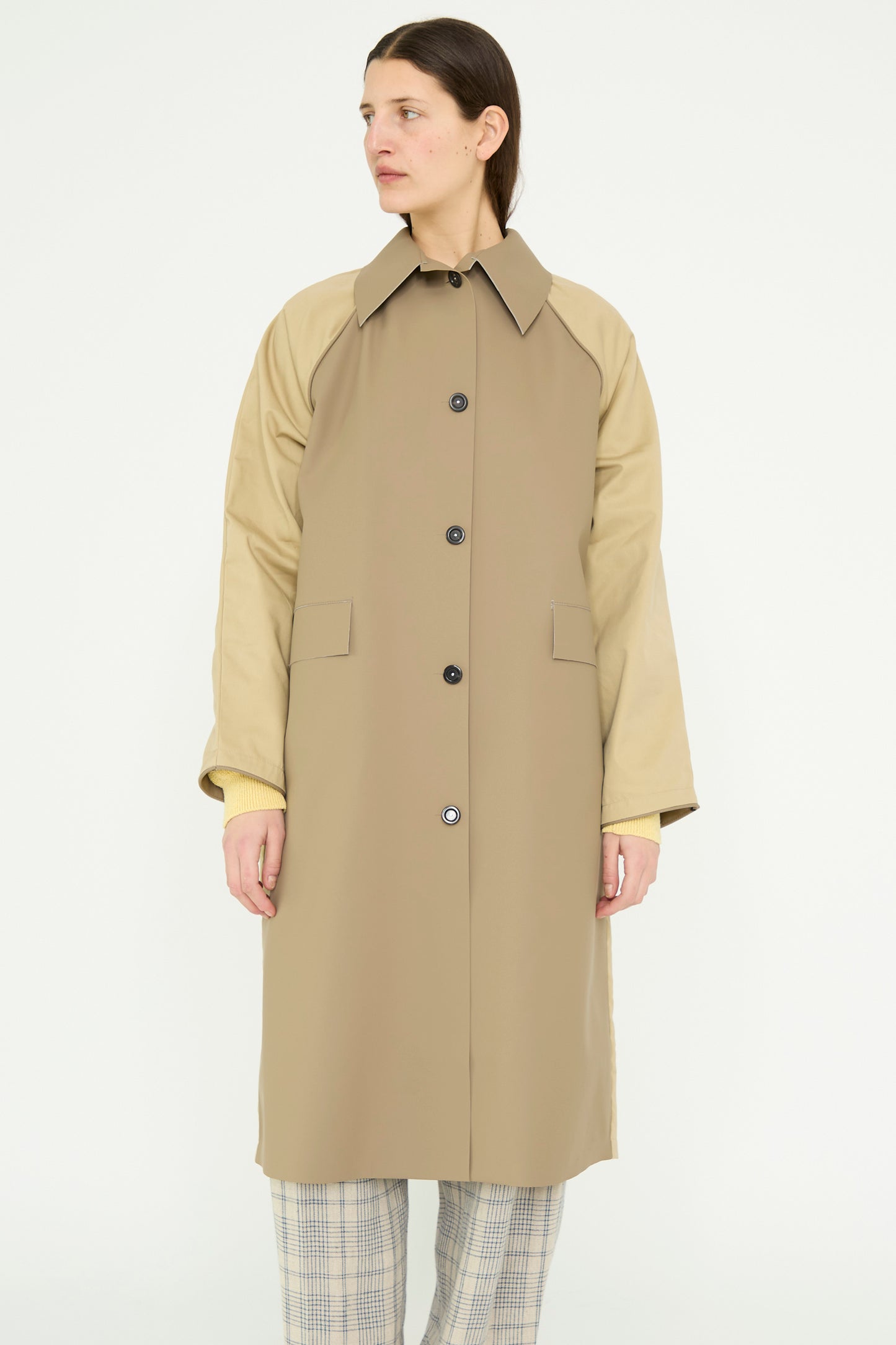 A person is styled in Kassl's Original Below Rubber Trench in Taupe and Beige, worn over a yellow top with plaid pants, highlighting its stylishly versatile polyester blend against a plain background.