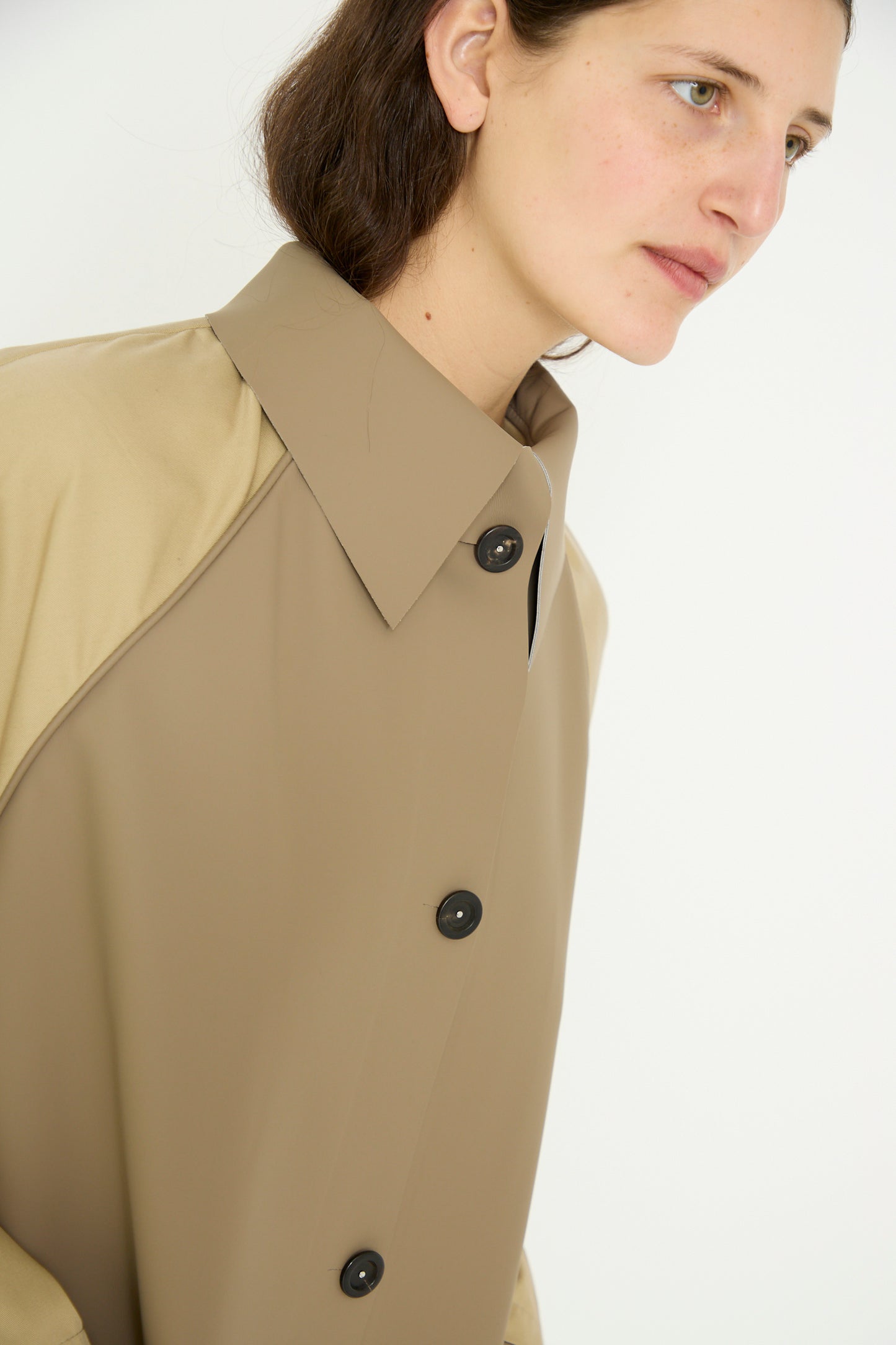 Clad in Kassl's Original Below Rubber Trench in taupe and beige, a person's gaze turns sideways against a plain background.