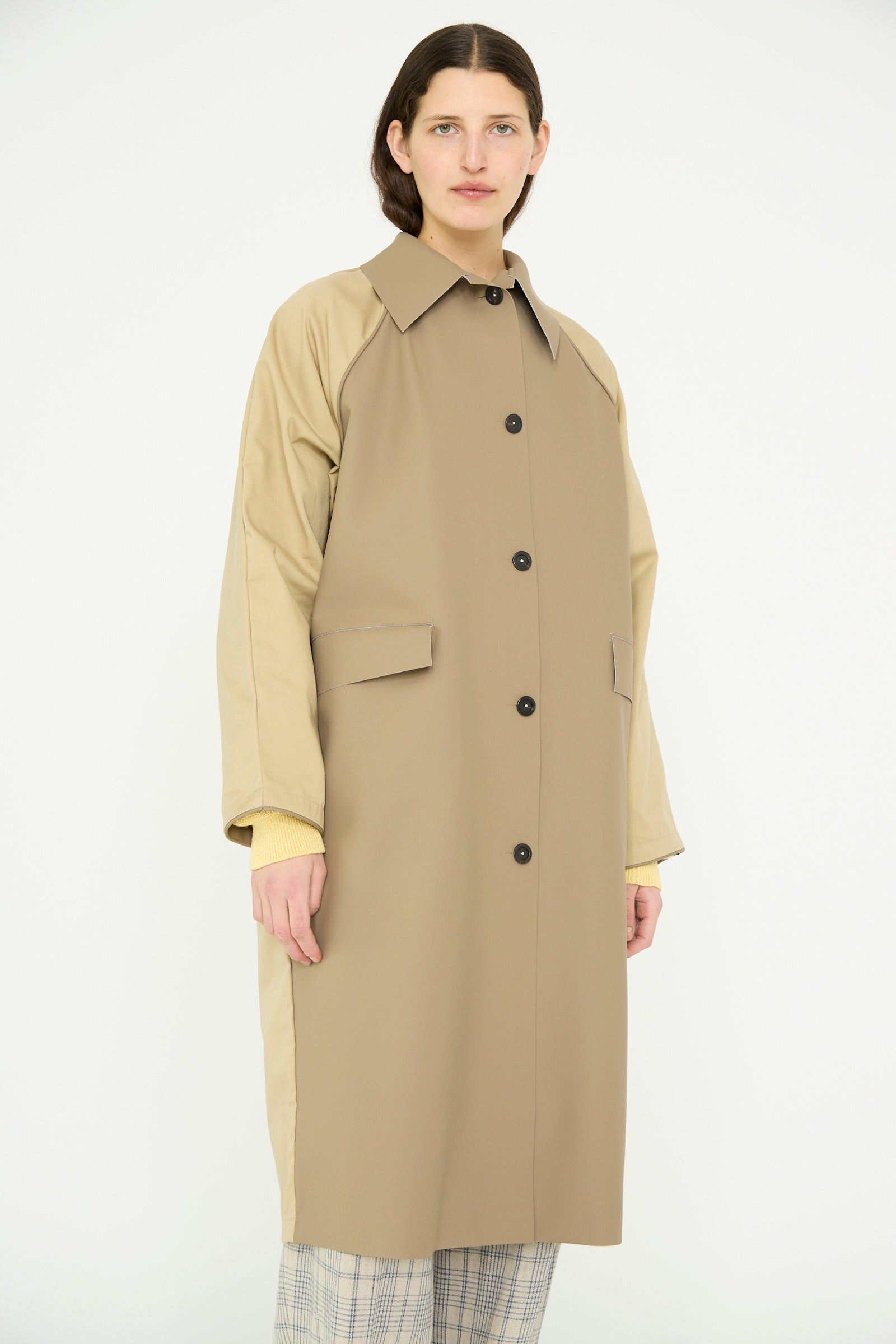 A person models the Kassl Original Below Rubber Trench in taupe and beige against a plain background. This stylish trench coat, made from a polyester blend, boasts buttons and pockets for both functionality and elegance.