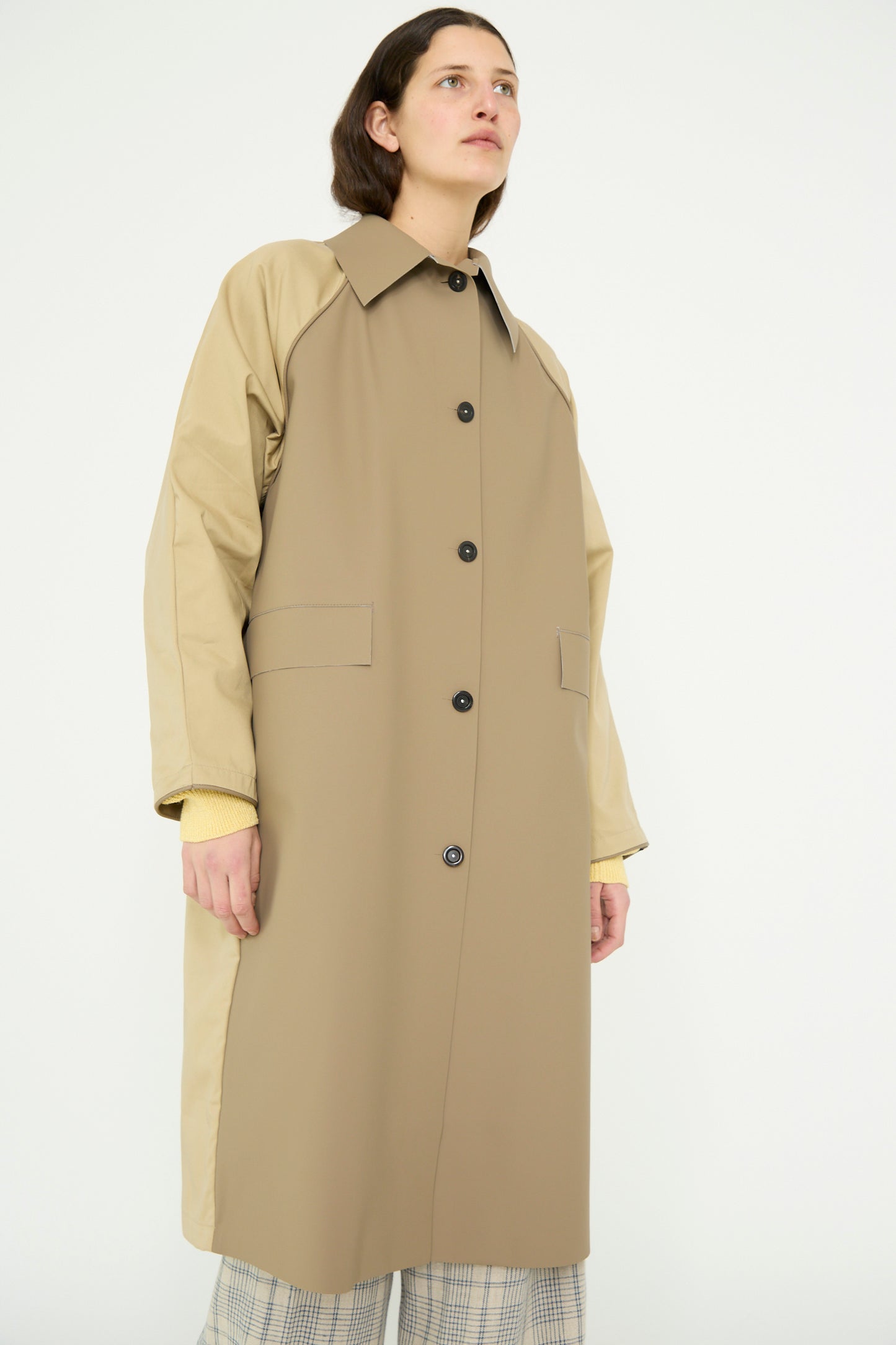 A person models Kassl's Original Below Rubber Trench in taupe and beige, a water-repellent polyester blend with a collared neckline and black buttons, against a plain background.