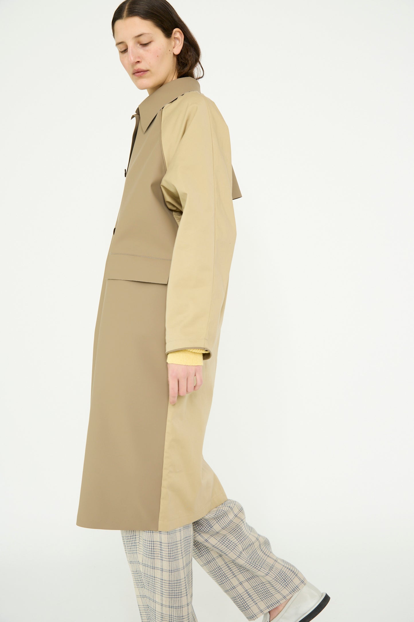 A person wearing Kassl's Original Below Rubber Trench in taupe and beige, paired with plaid pants, expertly balances on one foot against a plain background.