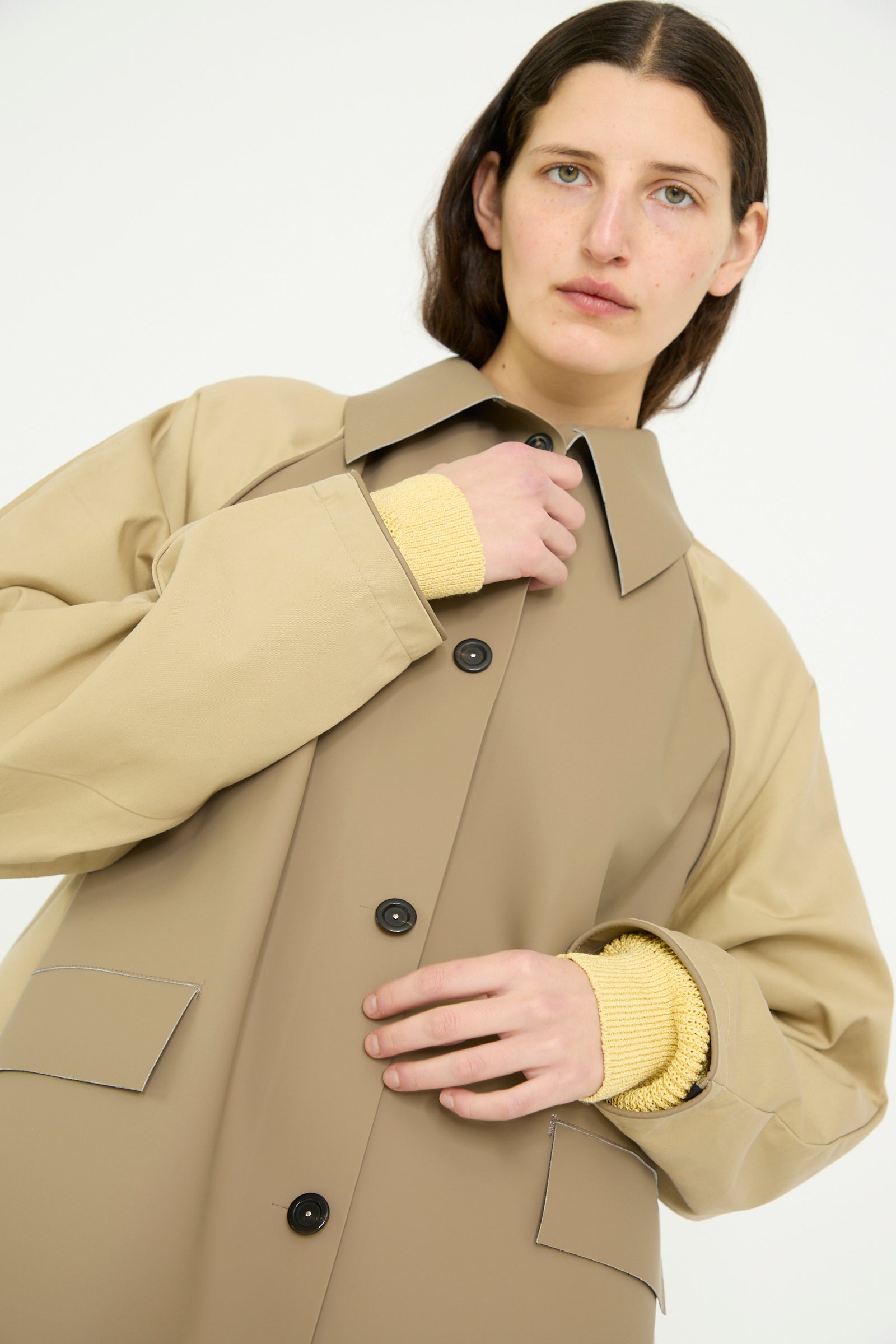 A person wearing Kassl's Original Below Rubber Trench in Taupe and Beige is standing against a light background. Their right hand reveals a yellow sweater sleeve beneath the polyester blend fabric.