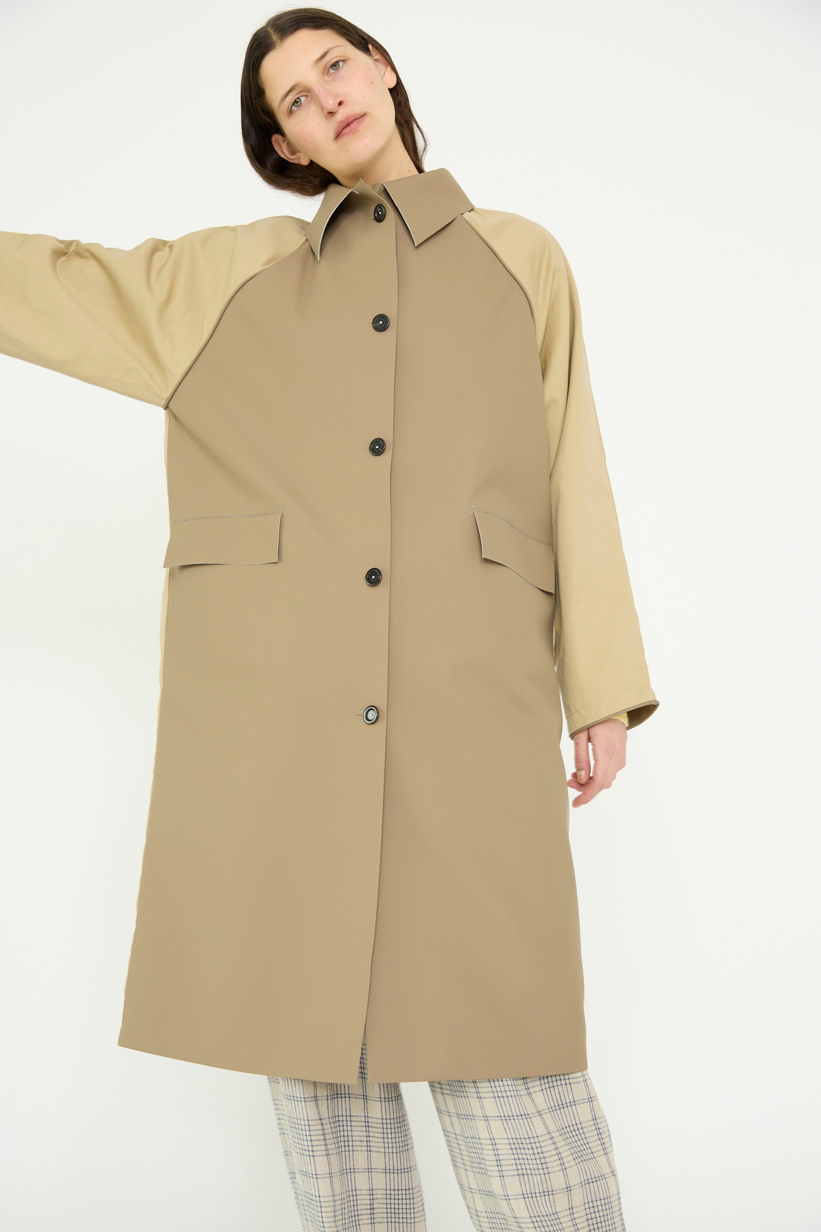 A person stands against a plain background wearing Kassl's Original Below Rubber Trench in taupe and beige, featuring button closure and beige sleeves crafted from a water-repellant polyester blend, paired with plaid pants.