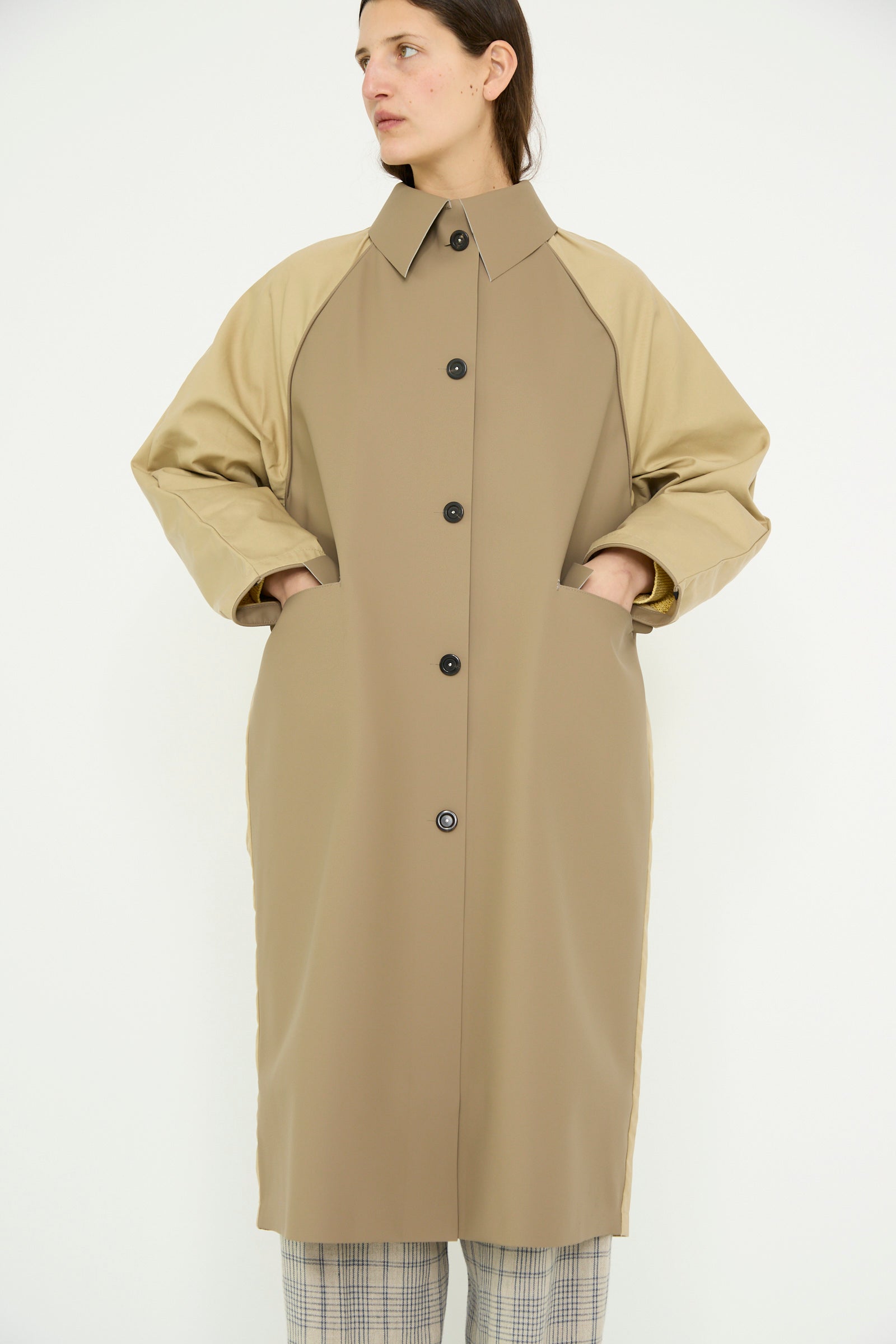 A person is wearing Kassl's Original Below Rubber Trench in Taupe and Beige, made of a water-repellent polyester blend with buttons done up and hands in pockets, paired with plaid pants, against a plain background.