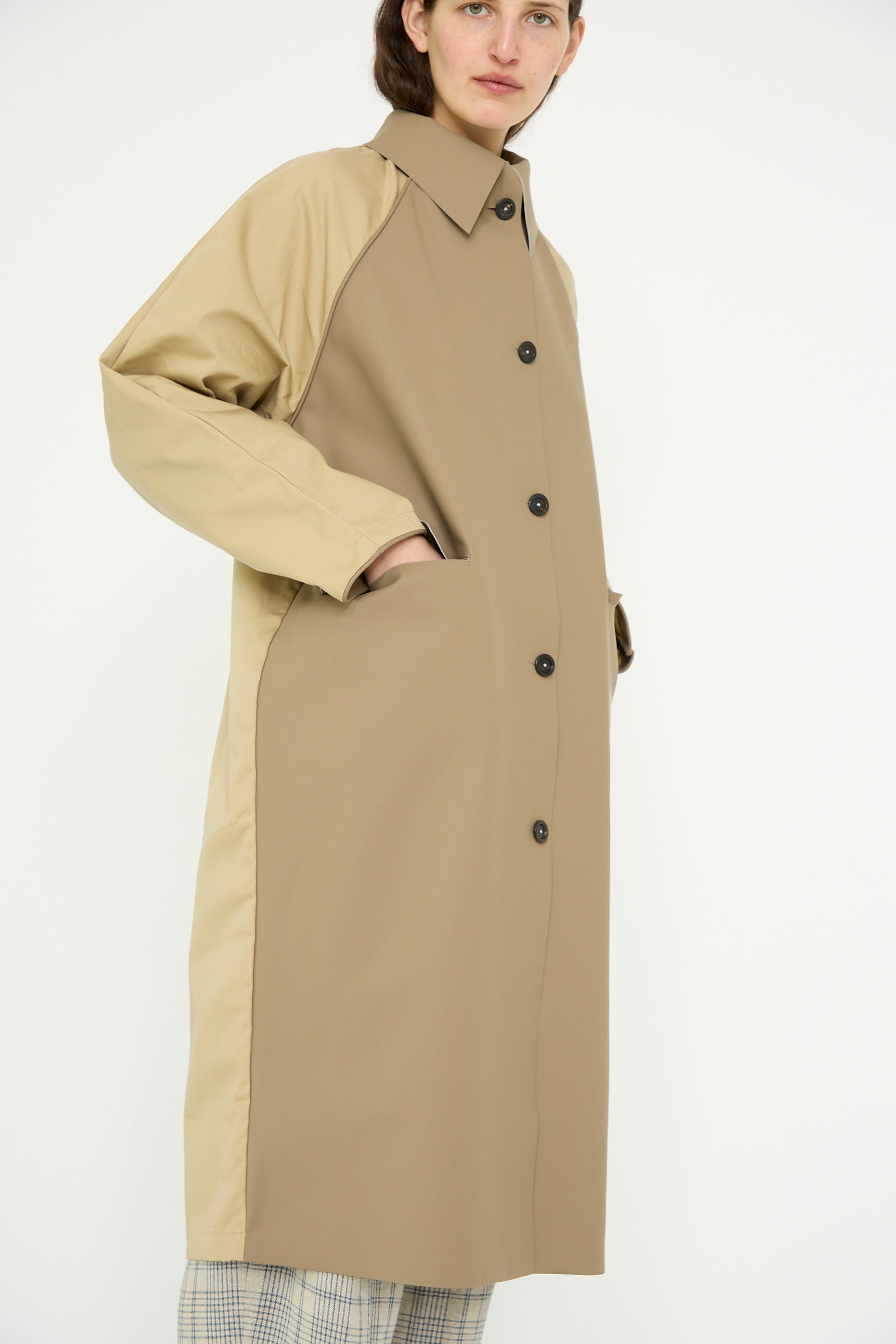 Someone poses against a white background wearing the Kassl Original Below Rubber Trench in taupe and beige, featuring black buttons, paired with plaid pants.