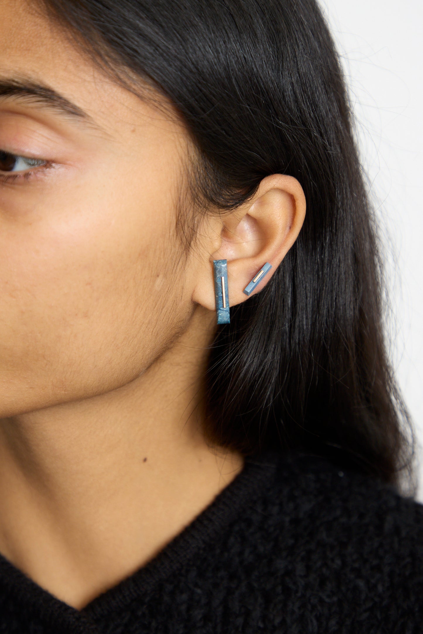 In a side view, a person wears Kathleen Whitaker's 14K Yellow Gold Long Staple Studs in Lapis, showcasing a blue geometric design accented by 14k gold. 