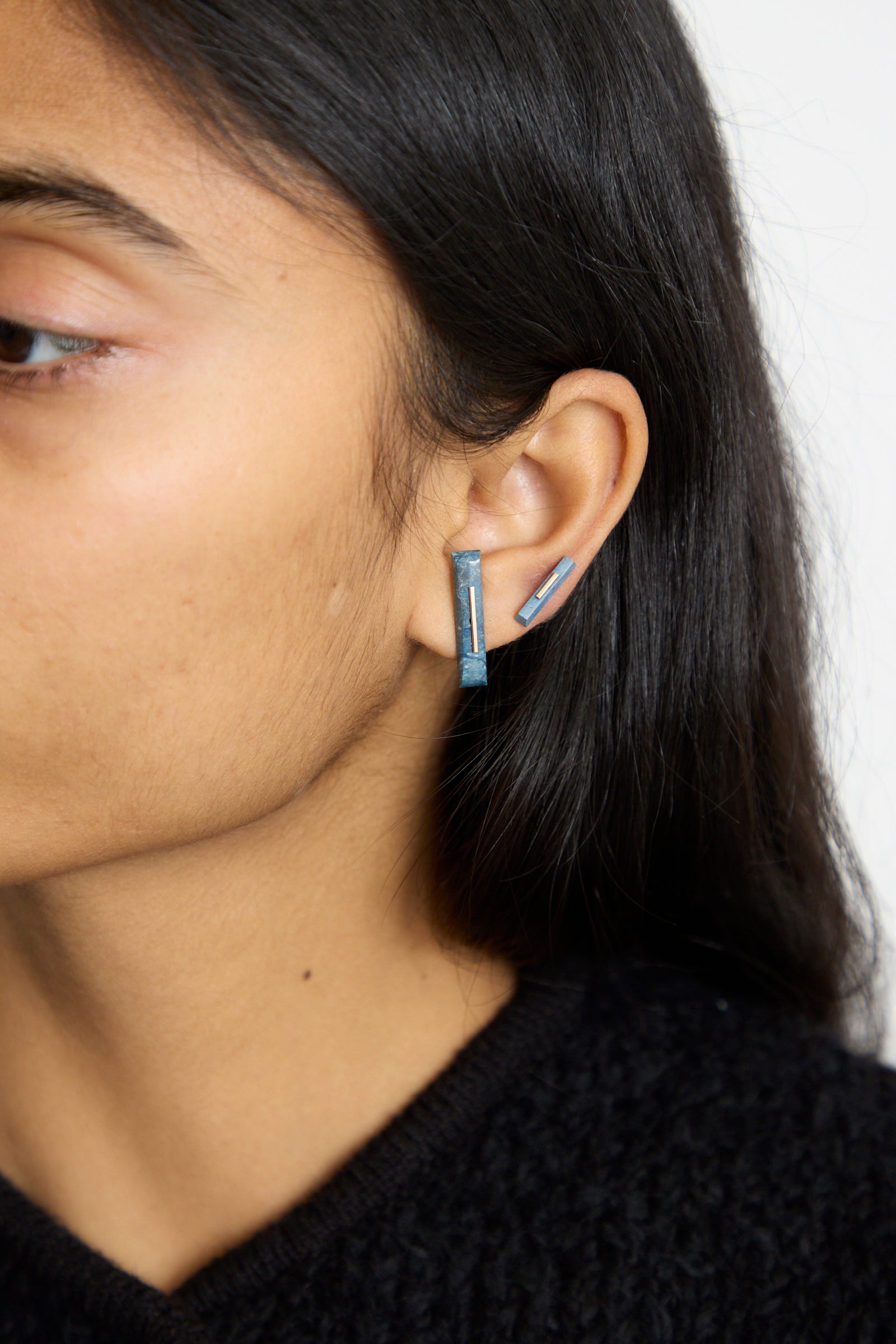 In a side view, a person wears Kathleen Whitaker's 14K Yellow Gold Long Staple Studs in Lapis, showcasing a blue geometric design accented by 14k gold. 