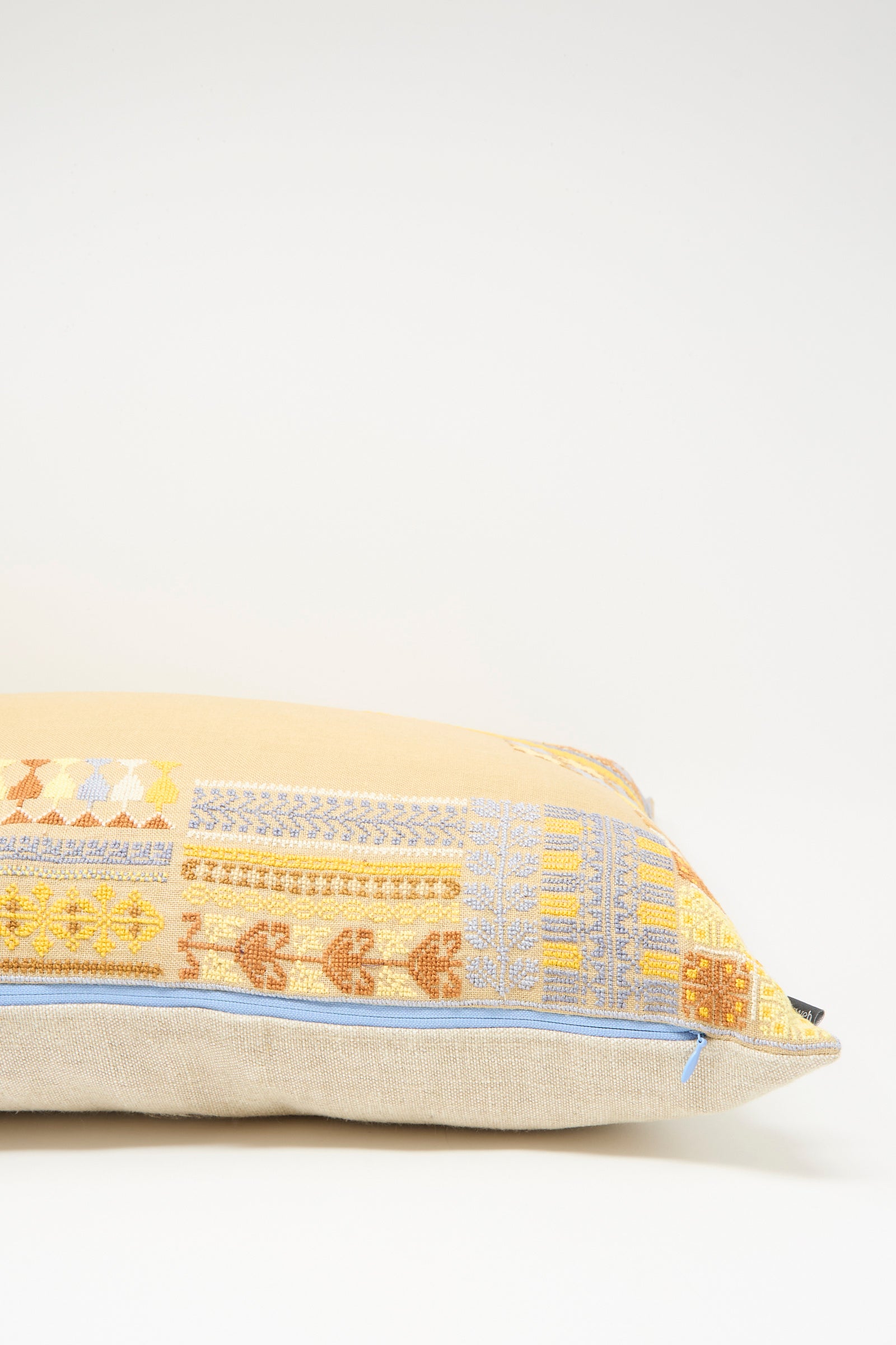 The Imm Bushra Hand Embroidered Pillow in Mustard by Kissweh features Belgian linen, a beige base, and hand-embroidered yellow, orange, and blue geometric patterns, finished with a light blue edge zipper.