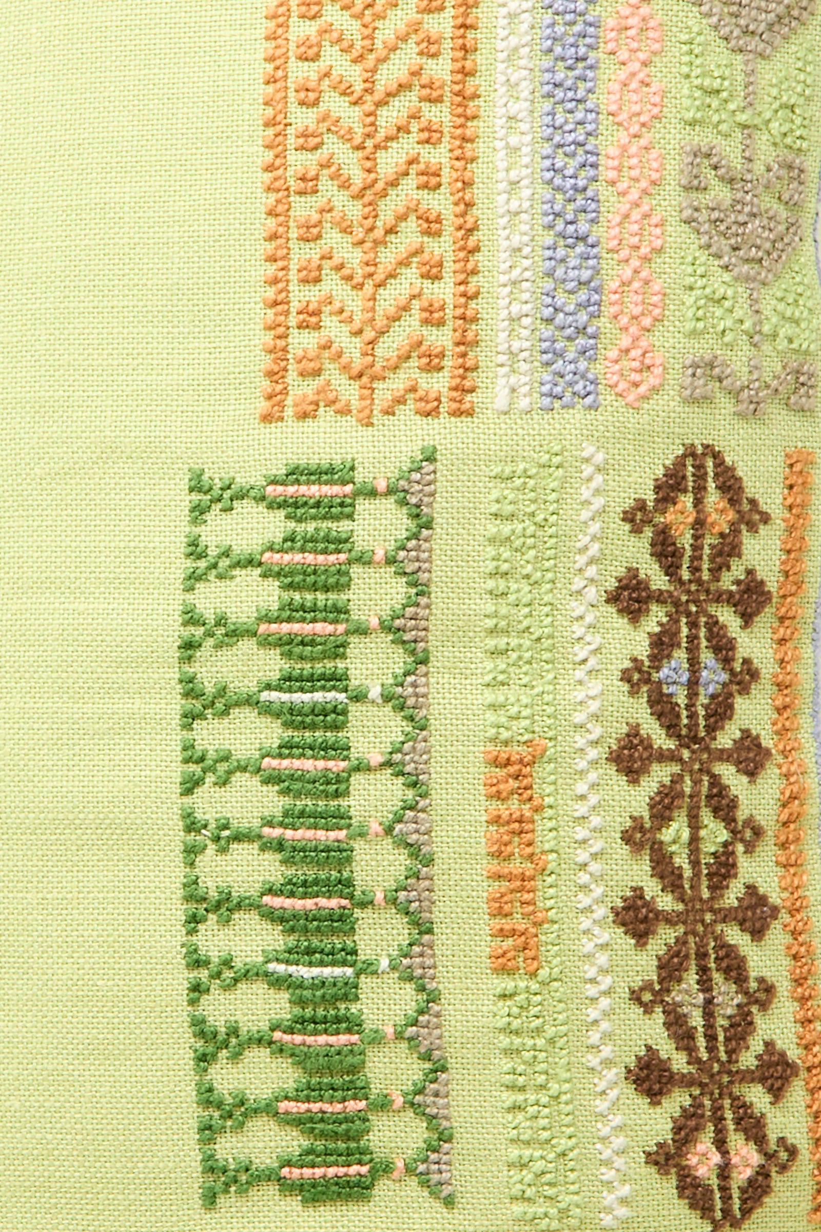 Close-up of geometric and floral patterns in green, orange, brown, lavender, and beige threads on light green fabric. Features the Kissweh Imm Bushra Hand Embroidered Pillow in Olive.