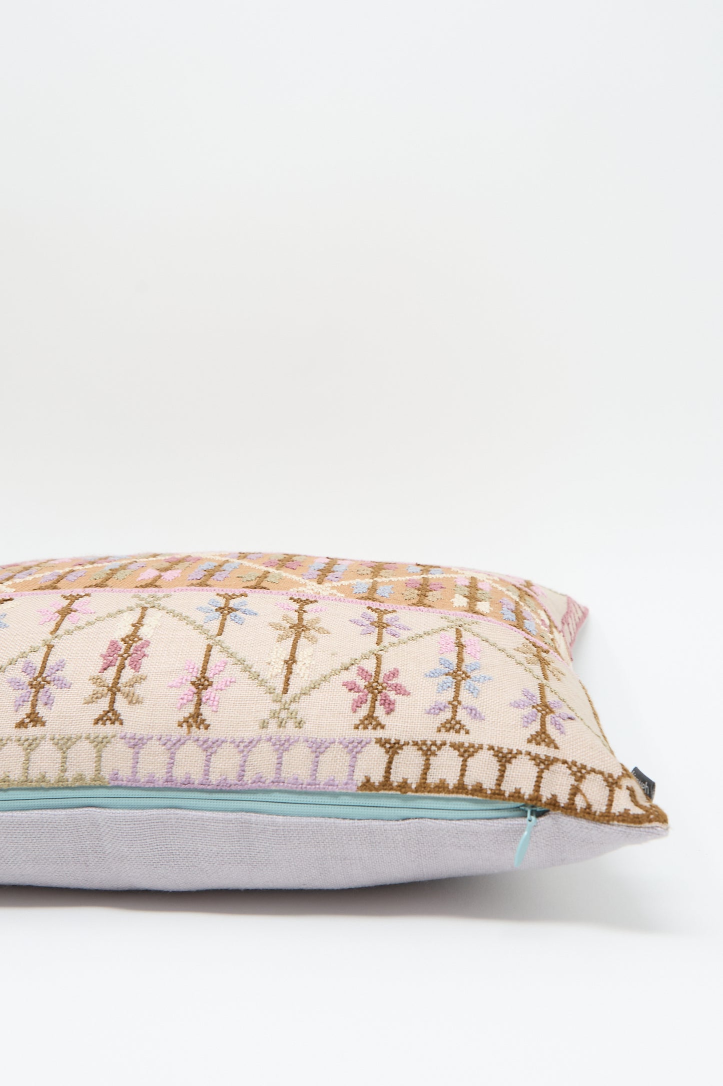 A rectangular decorative pillow with a detailed, multicolored woven pattern on the front, featuring various geometric shapes and lines set against a plain light background, complemented by luxurious Belgian linen and a plush down feather insert—Kissweh's Imm Omar Hand Embroidered Pillow in Beige and Cognac.