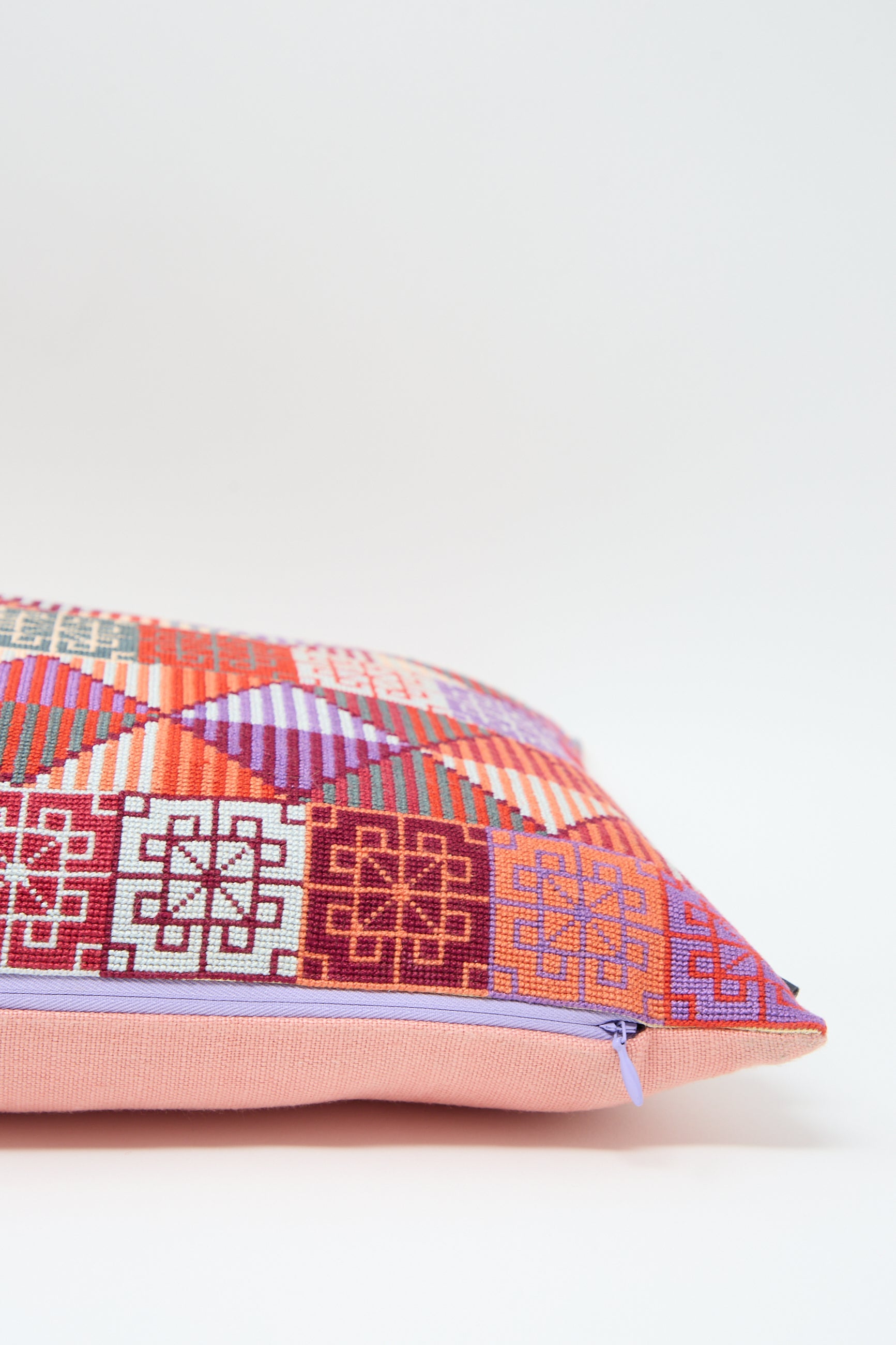 A vibrant, patterned cushion with geometric hand embroidery in various shades of red, pink, orange, and purple, featuring a light pink zipper along one edge against a plain white background. Introducing the Ola Hand Embroidered Pillow in Crimson by Kissweh.