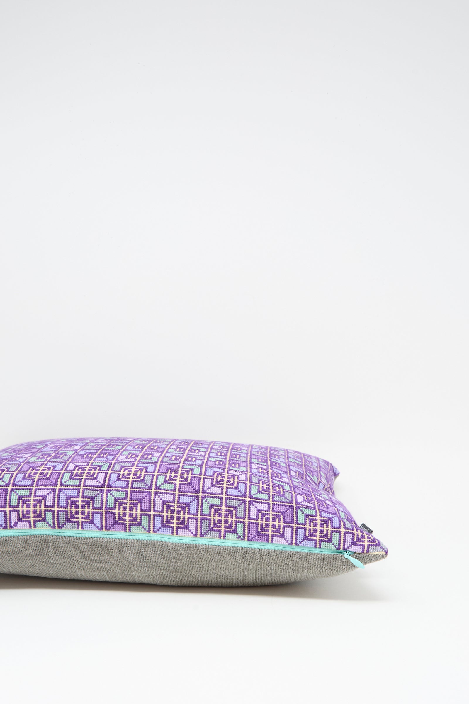 The Kissweh Rana Hand Embroidered Pillow in Lilac and Purple I, featuring a geometric design, rests on a light surface against a white background.