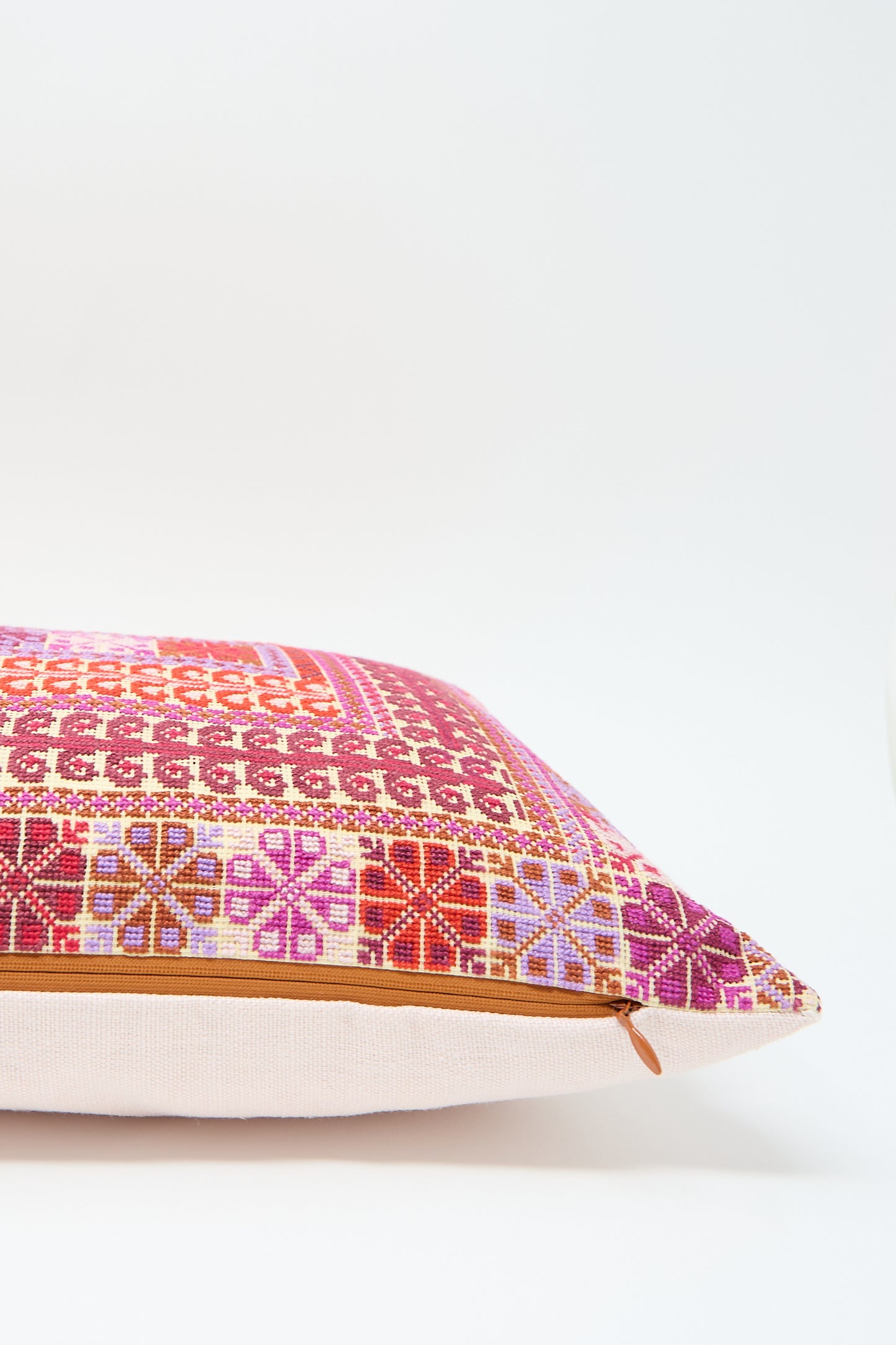 The Kissweh Yasmin Hand Embroidered Pillow in Crimson is a decorative lumbar pillow featuring a geometric, multi-colored pattern in shades of red, pink, and orange, crafted from luxurious Belgian linen with intricate hand embroidery on a beige base and a visible zipper on the side.