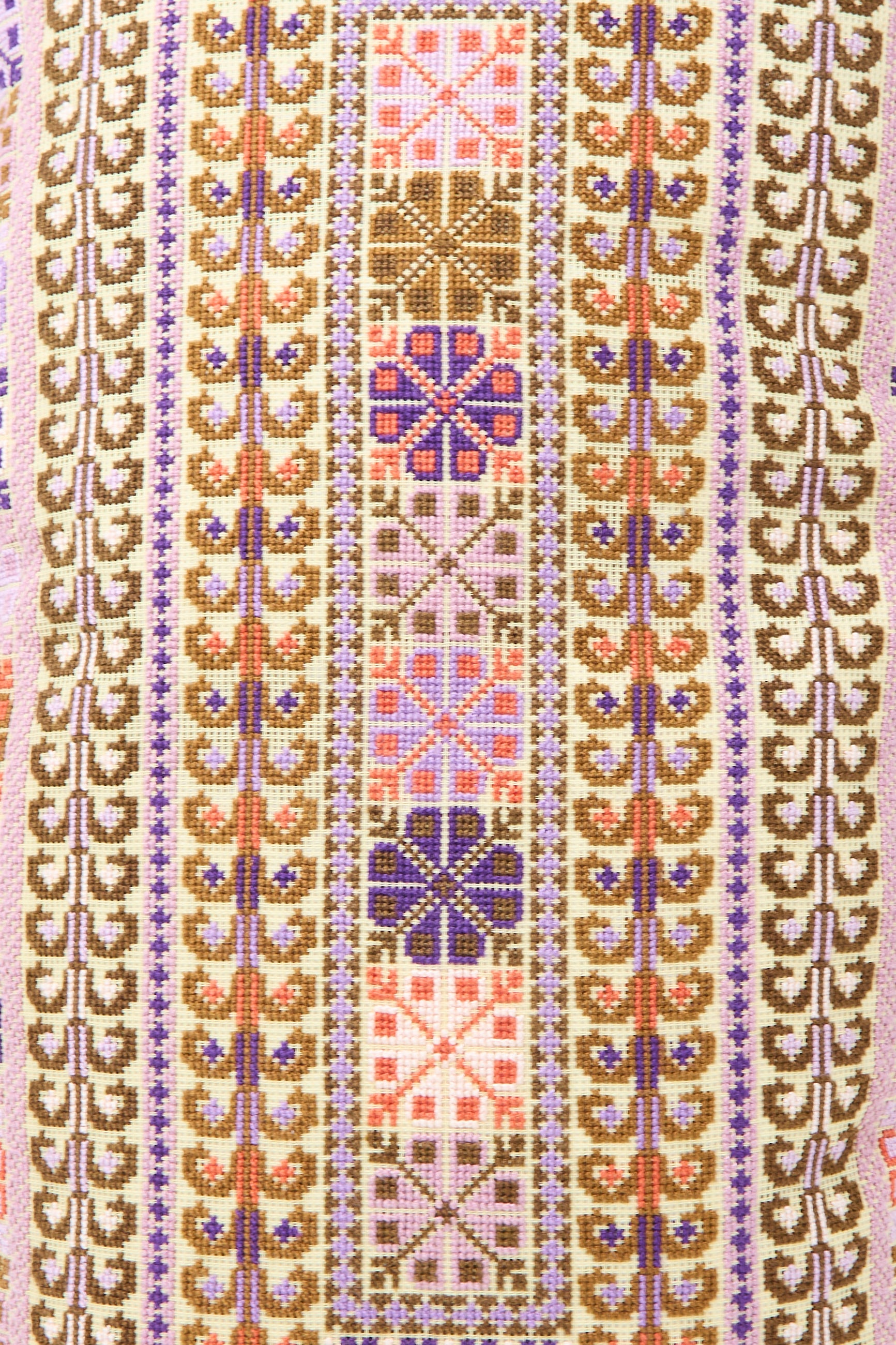 A close-up of a patterned textile with vertical rows of geometric hand embroidery in pink, brown, purple, and orange colors on the Kissweh Yasmin Hand Embroidered Pillow in Lilac and Coral.