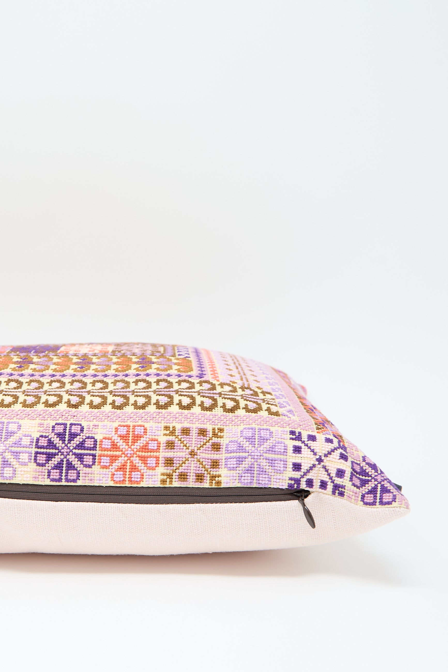 The Yasmin Hand Embroidered Pillow in Lilac and Coral from Kissweh is a rectangular lumbar pillow with a colorful geometric pattern featuring shades of pink, purple, and orange on a white background. Crafted from Libeco Belgian linen, the cushion showcases exquisite geometric hand embroidery and has a zipper on one side.