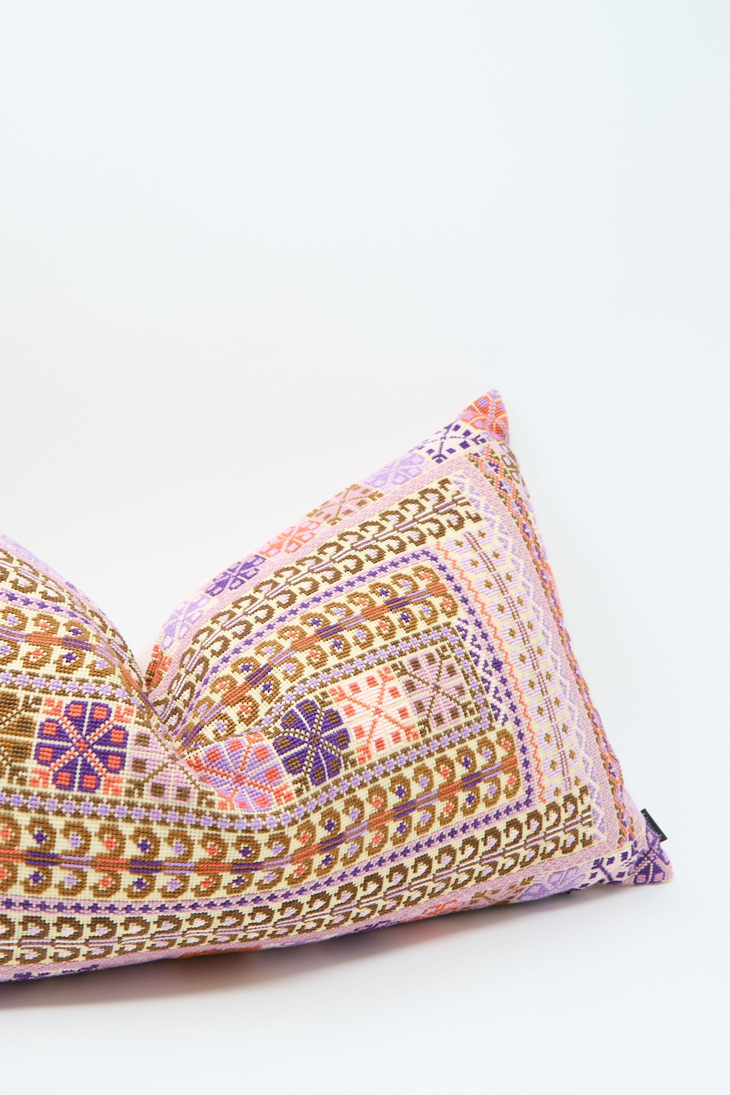 A rectangular lumbar pillow with a colorful, intricate geometric pattern in shades of purple, pink, and brown is displayed against a plain white background. The detailed geometric hand embroidery adds an elegant touch to the piece. This is the Yasmin Hand Embroidered Pillow in Lilac and Coral by Kissweh.