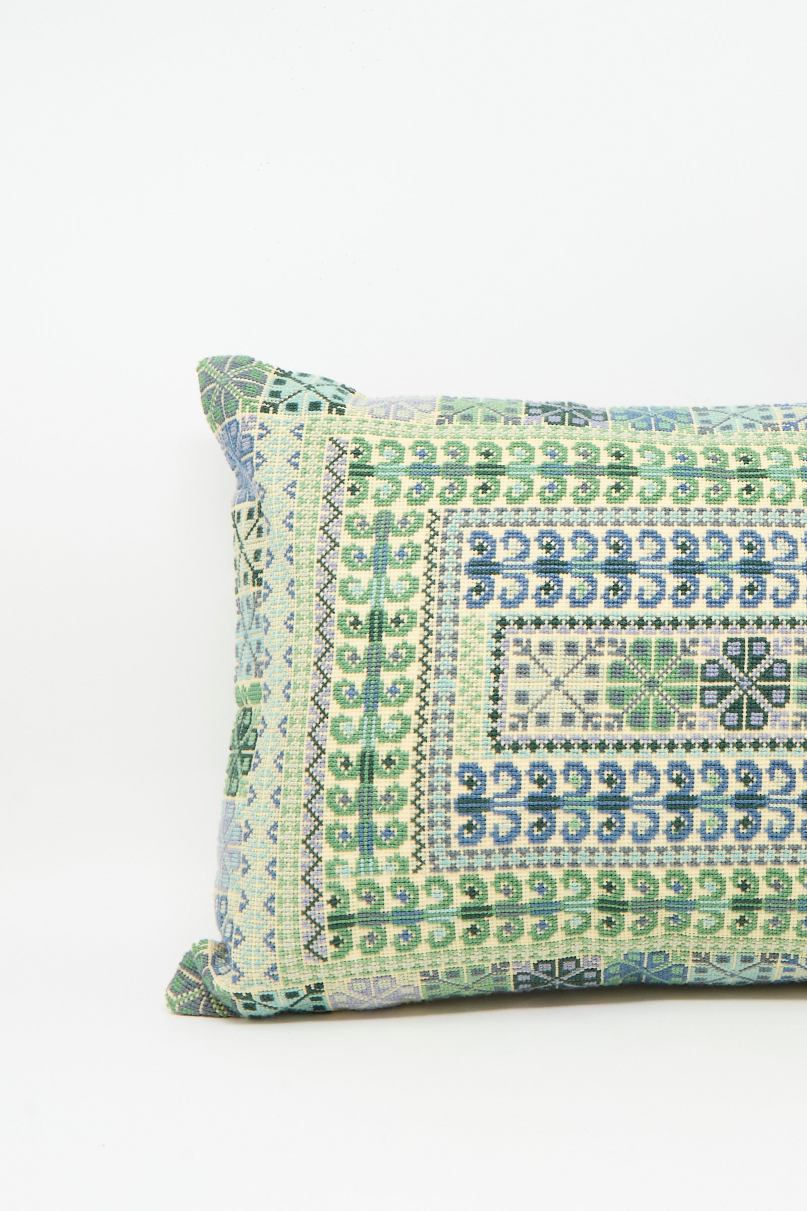 The Yasmin Hand Embroidered Pillow in Olive by Kissweh features a geometric design in green, blue, and black on Belgian linen with a white background. It is shown resting against a plain white surface.