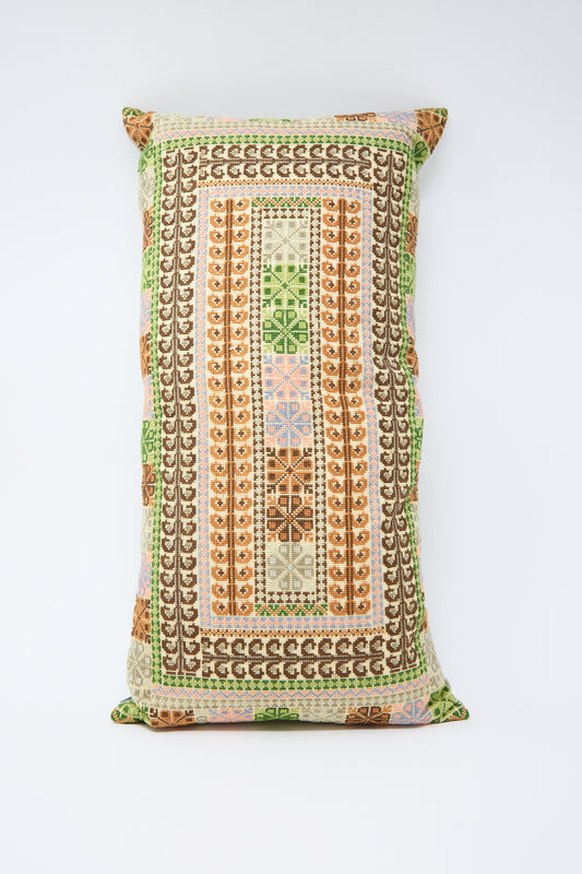 A Yasmin Hand Embroidered Pillow in Olive and Slate from Kissweh adorned with a detailed geometric pattern in green, brown, orange, and cream colors against a white Belgian linen background.