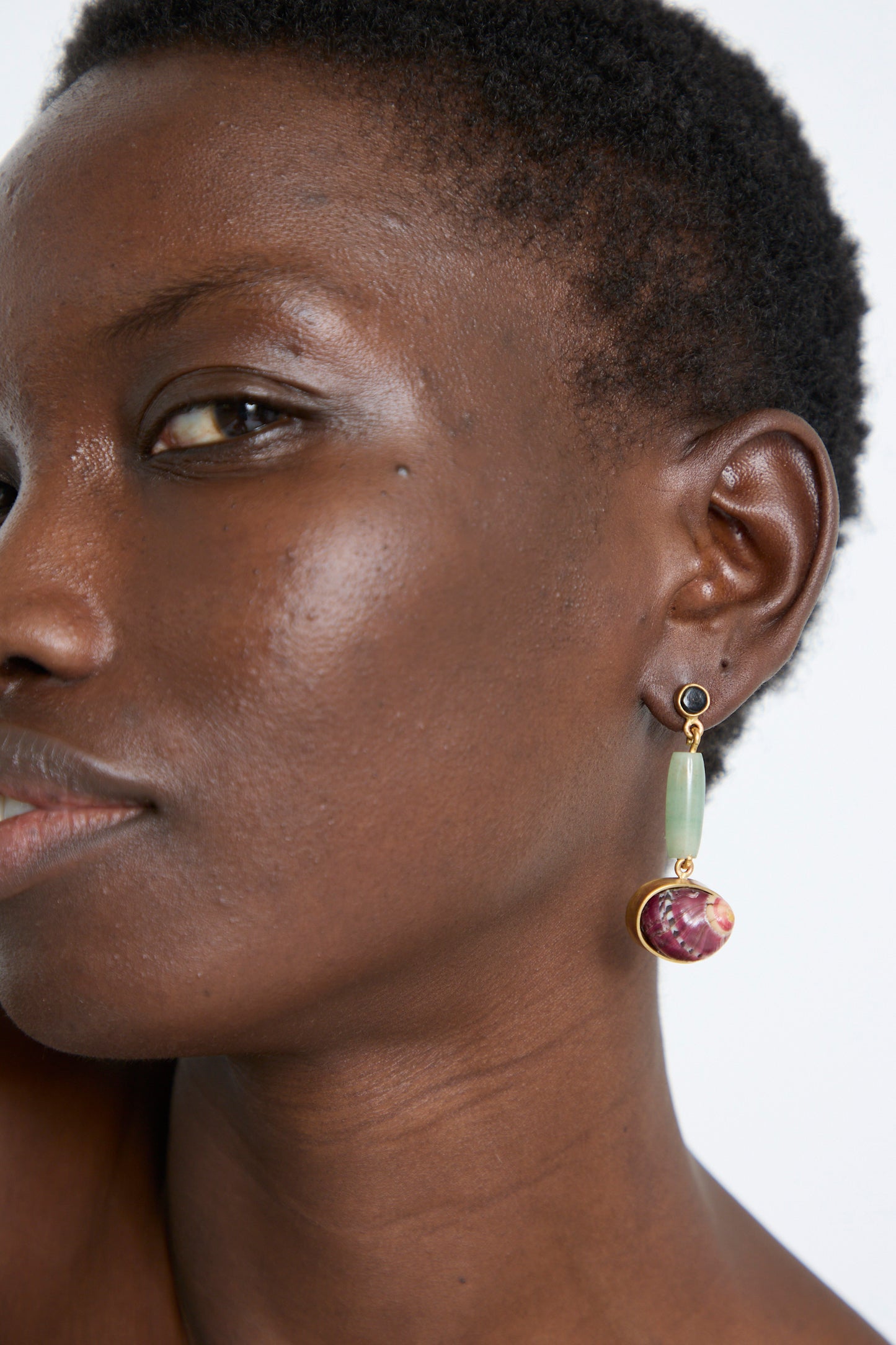 Close-up of a person wearing La Ma r's Sterling Silver No. 09 Earrings in Green and Red I, featuring a green oval and a pink stone, delicately set in sterling silver. The background is neutral.