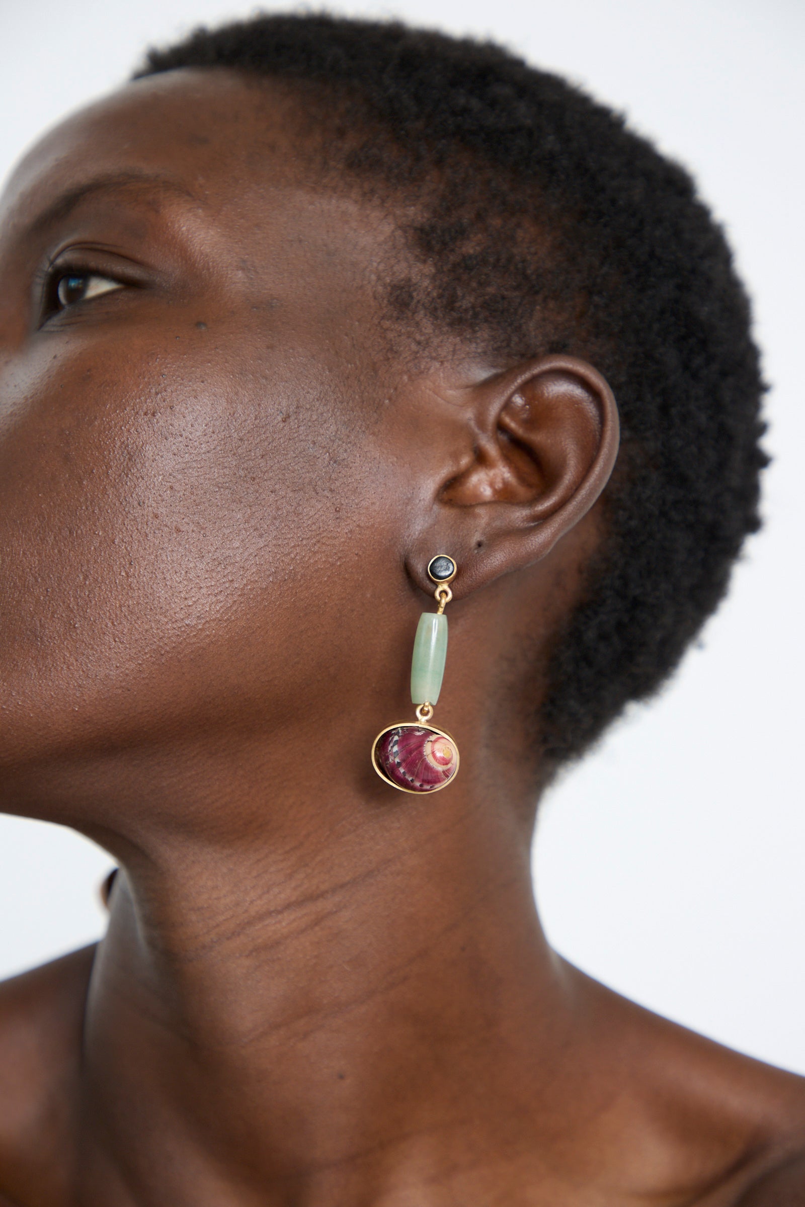 Profile view of a person with short hair wearing the Sterling Silver No. 09 Earrings in Green and Red I by La Ma r, which feature a green bead, a red textured stone, and a delicate spiral shell, all beautifully crafted in sterling silver.