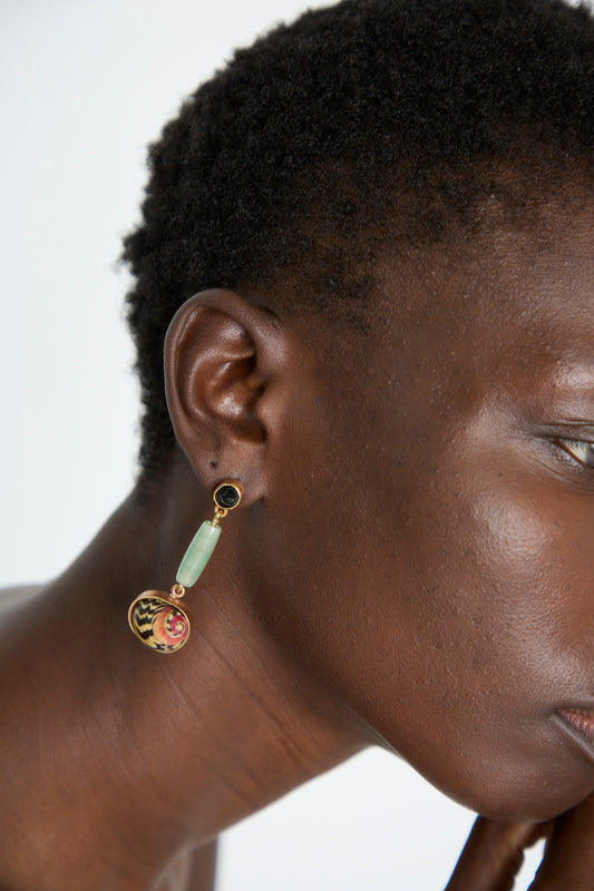 A person wearing the La Ma r Sterling Silver No. 09 Earrings in Green and Red I, which feature a sterling silver base, a green rectangular stone, a small black round stone, and a colorful circular pendant.