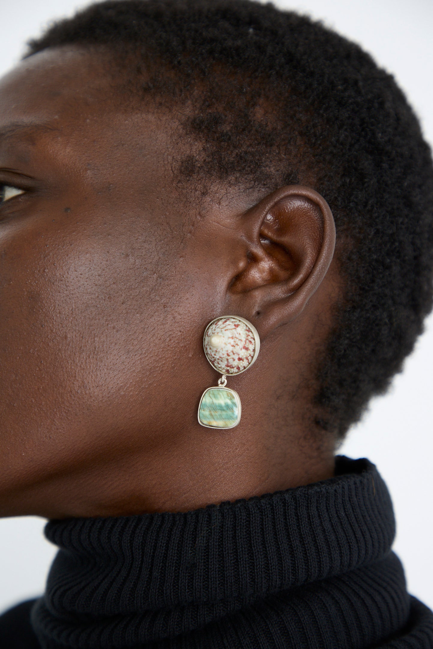 A close-up of a person in a black turtleneck wearing the Sterling Silver No. 09 Earring by La Mar, featuring a circular top and a rectangular green pendant.