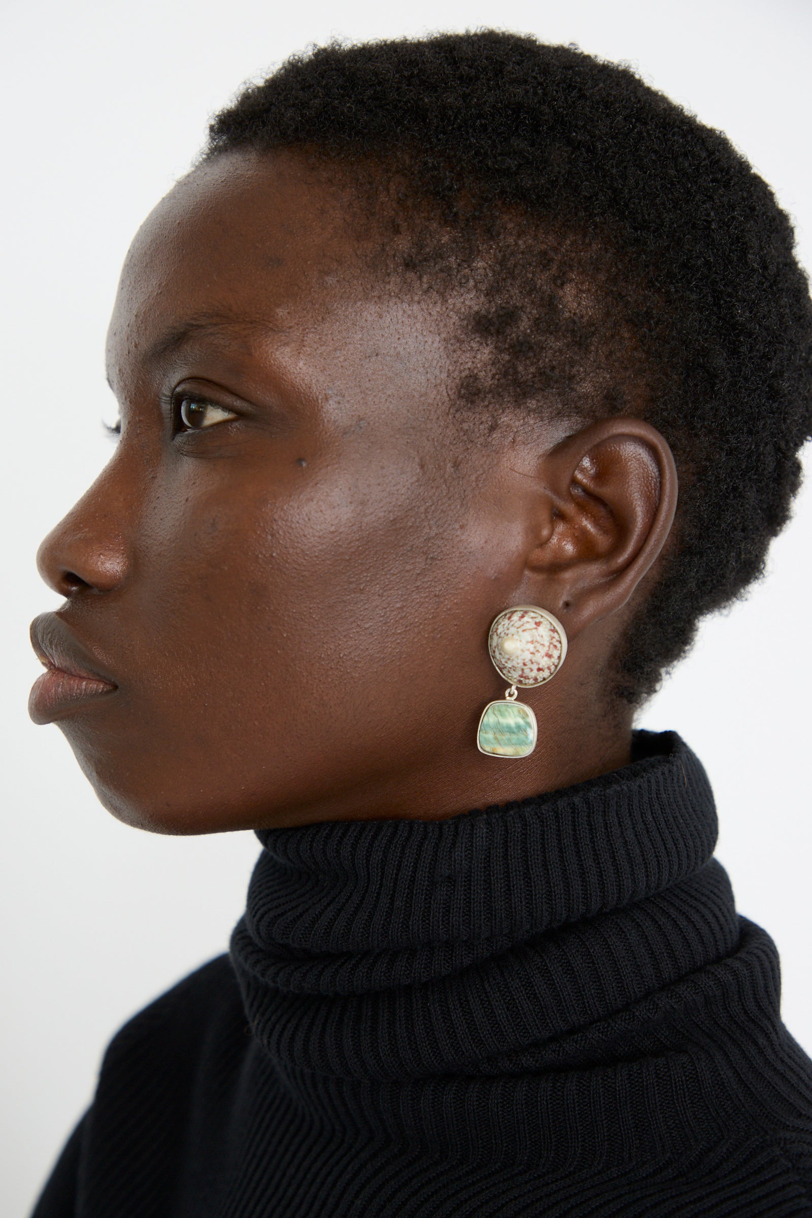 Side profile of a person wearing a black turtleneck, and the La Ma r's Sterling Silver No. 09 Earrings in Green and Pink Shell, set against a white background.