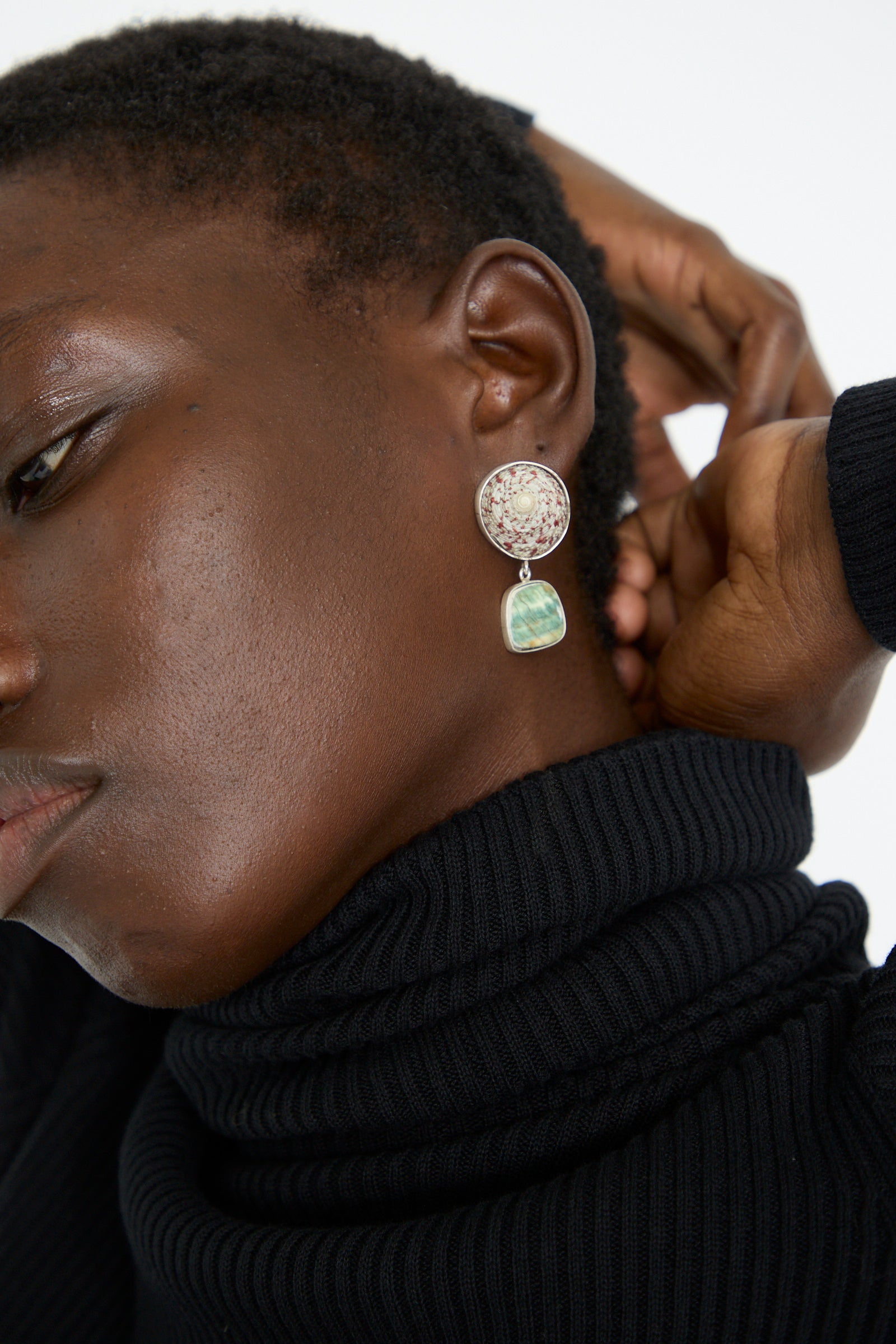A person wearing a black turtleneck and La Mar's Sterling Silver No. 09 Earrings in Green and Pink Shell, with a hand raised to their head.