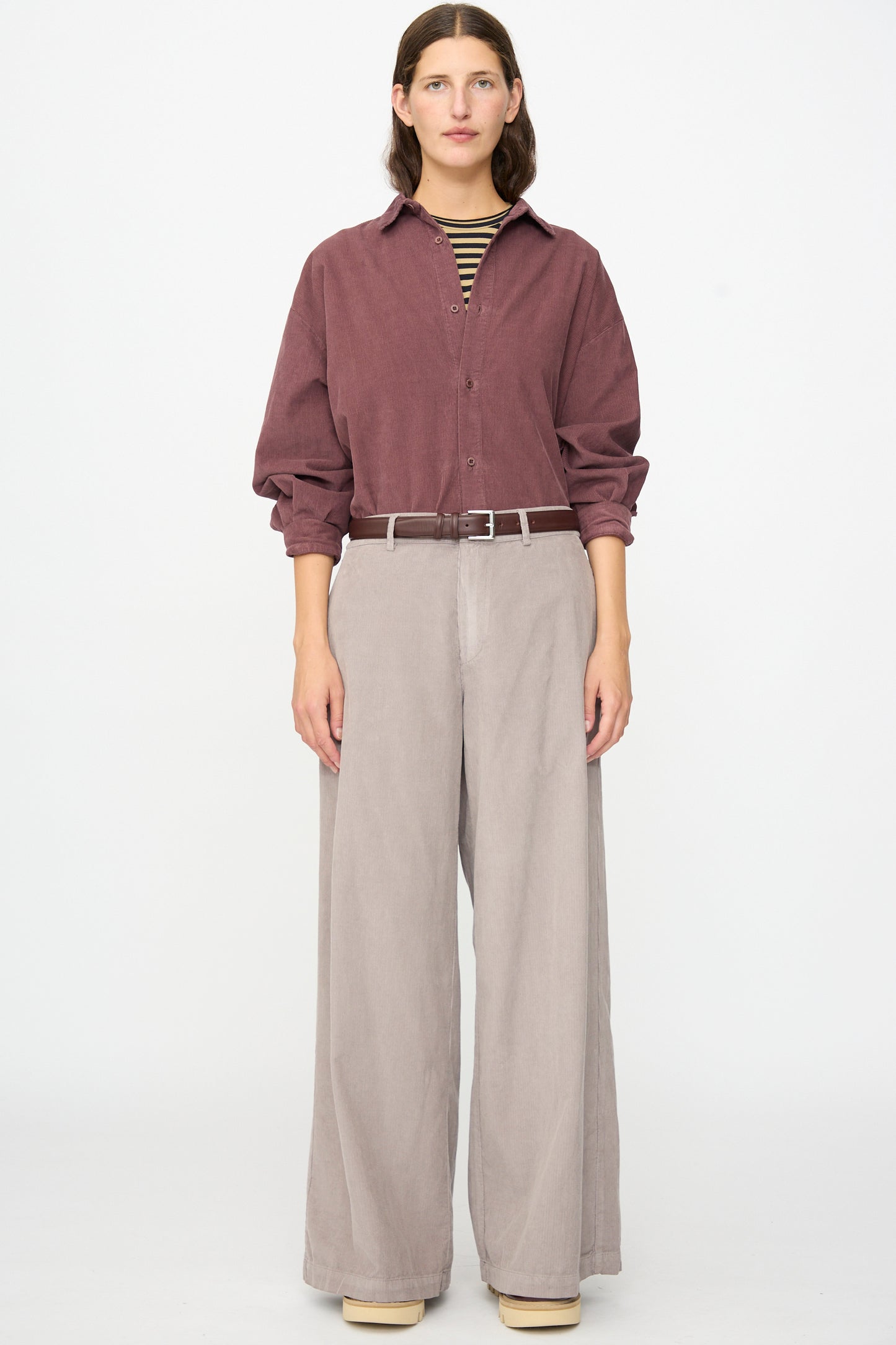 A person stands against a white background, wearing a maroon button-up shirt, striped undershirt, and beige wide-leg Labo.Art Binocchio Trouser in Malva crafted from soft cotton needlecord, secured with a belt, paired with beige shoes.