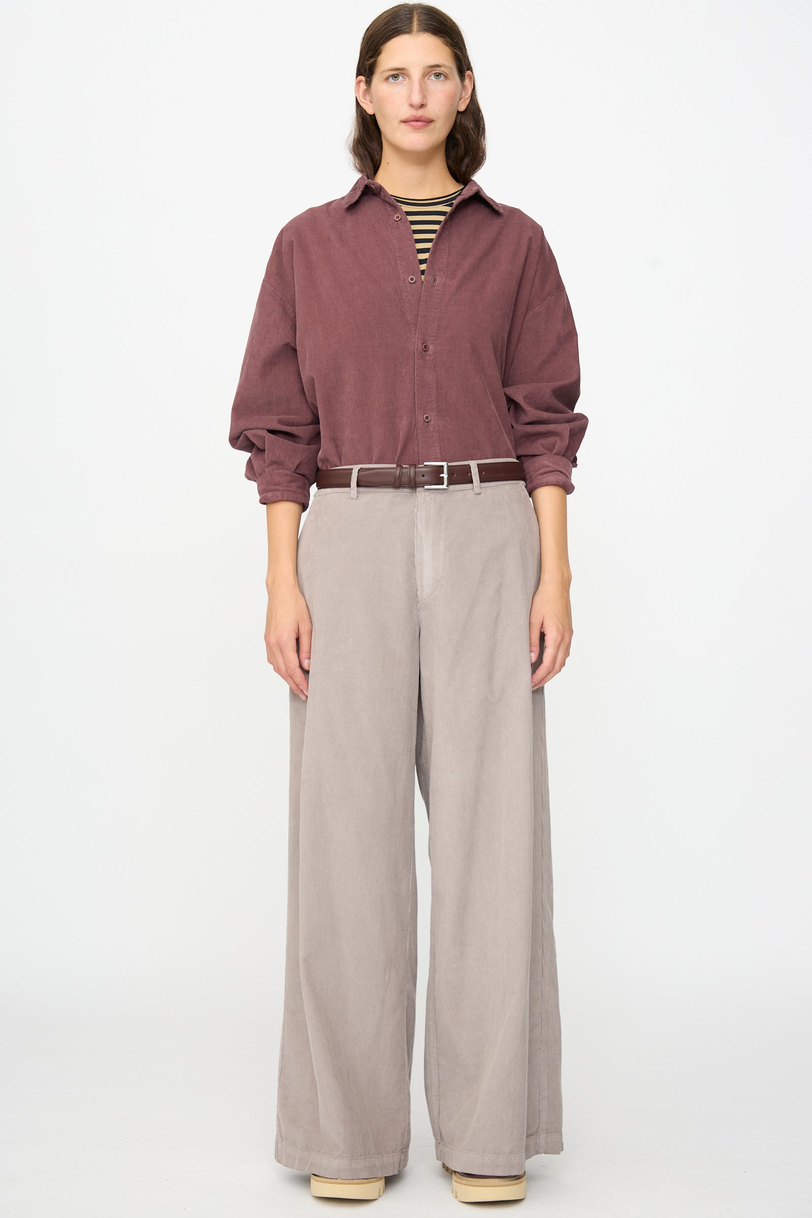 A person stands against a white background, wearing a maroon button-up shirt, striped undershirt, and beige wide-leg Labo.Art Binocchio Trouser in Malva crafted from soft cotton needlecord, secured with a belt, paired with beige shoes.