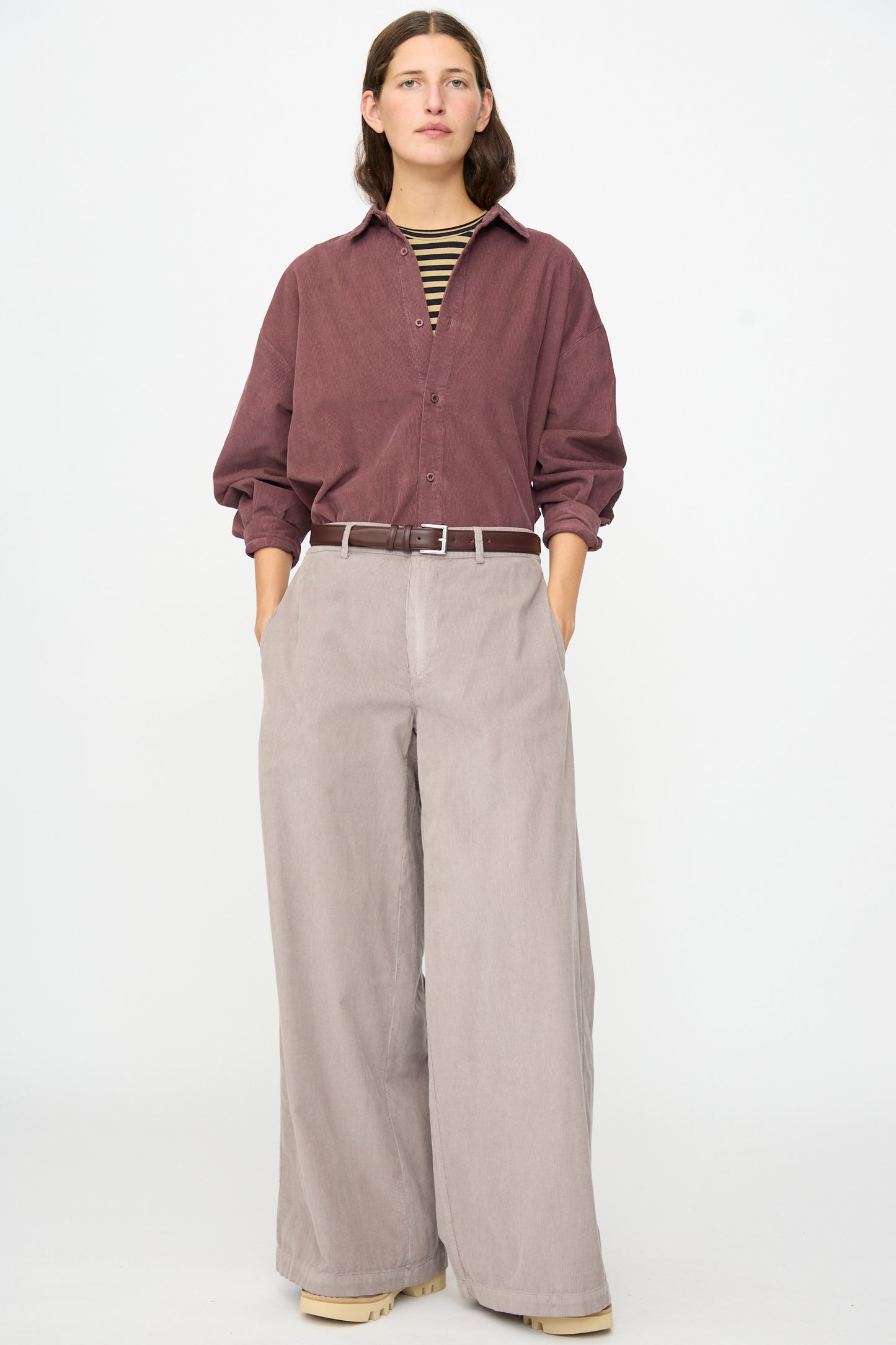 A person stands against a plain background wearing the Labo.Art Binocchio Trouser in Malva—wide-leg beige pants crafted from soft cotton needlecord—paired with a maroon button-up shirt, striped undershirt, beige shoes, and hands in pockets.