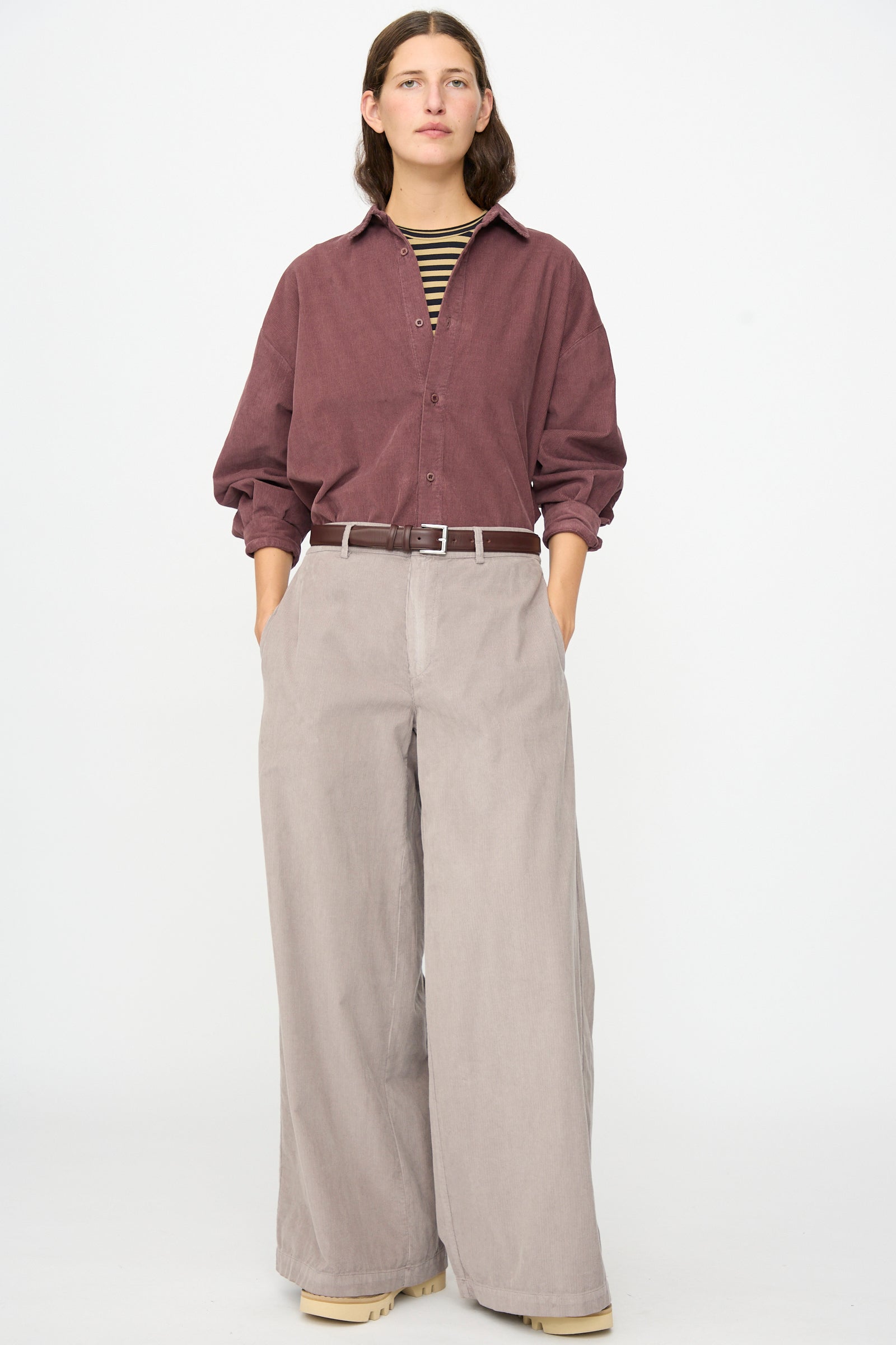 A person stands against a plain background wearing the Labo.Art Binocchio Trouser in Malva—wide-leg beige pants crafted from soft cotton needlecord—paired with a maroon button-up shirt, striped undershirt, beige shoes, and hands in pockets.