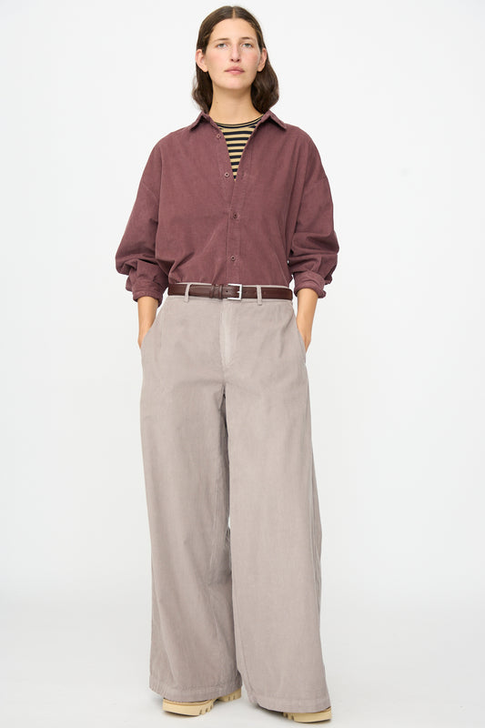 A person stands against a plain background wearing the Labo.Art Binocchio Trouser in Malva—wide-leg beige pants crafted from soft cotton needlecord—paired with a maroon button-up shirt, striped undershirt, beige shoes, and hands in pockets.