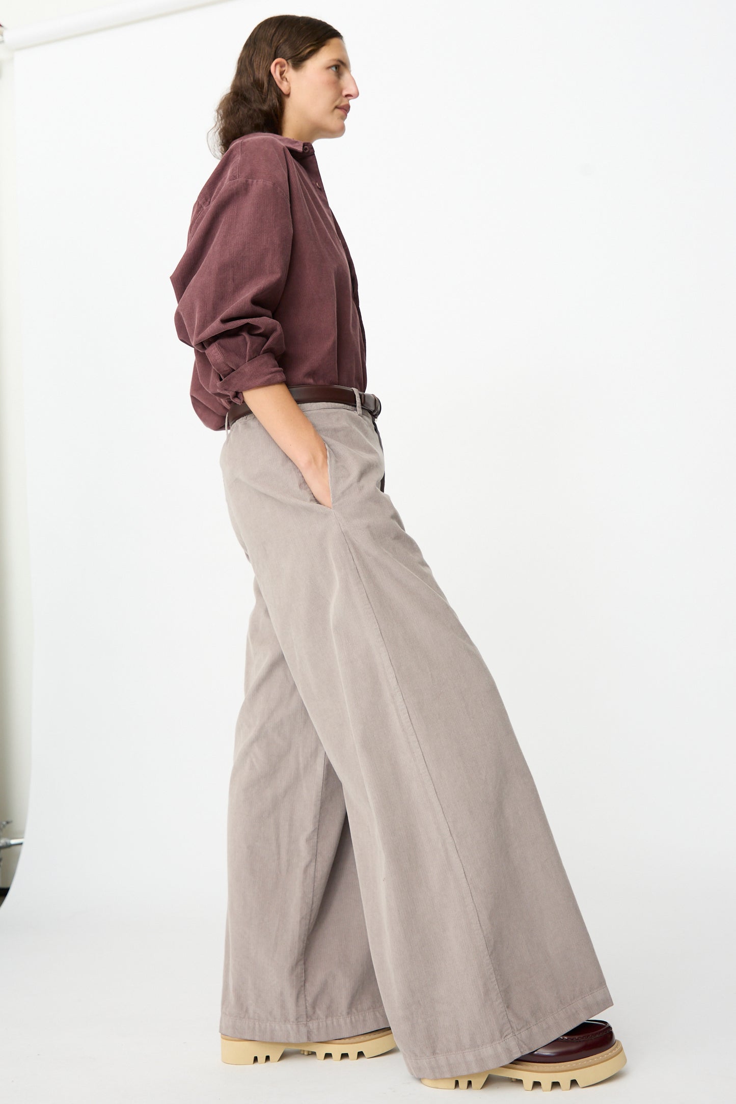 A person stands in profile against a white background, wearing the Labo.Art Binocchio Trouser in Malva—a wide-leg pant crafted from soft cotton needlecord—paired with a burgundy shirt, platform shoes, and hands casually tucked in pockets.